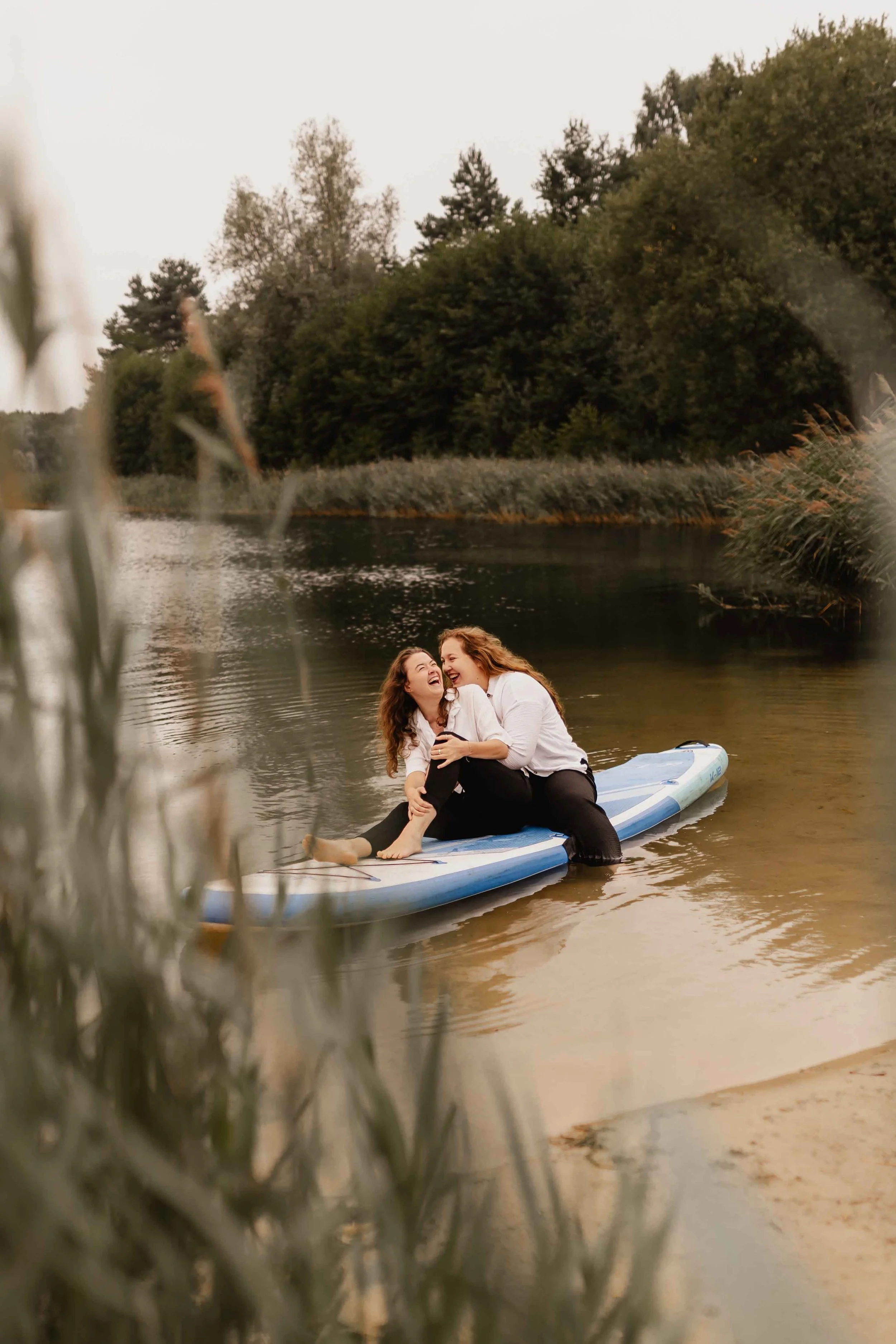 Zwei Frauen sitzen umarmt und lachend auf einem SUP-Board am See, eingerahmt von Schilf und Bäumen