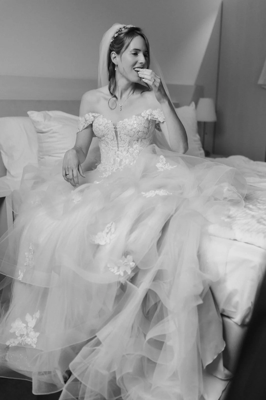 Schwarz-Weiß-Fotografie einer lachenden Braut im romantischen Spitzenbrautkleid mit Tüllrock und Schleier, die beim Getting Ready entspannt auf dem Bett im Heide Hotel Reinstorf bei Lüneburg sitzt.