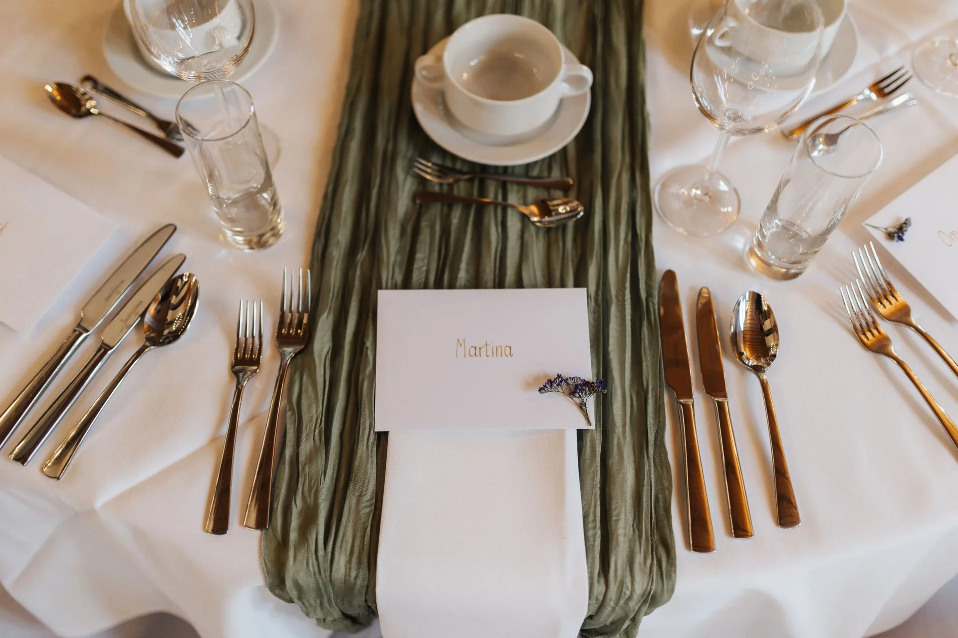 Elegante Tischdekoration bei einer Hochzeit im Heide Hotel Reinstorf mit grüner Stoffserviette, Namenskarte, goldenem Besteck und weißer Tischdecke.