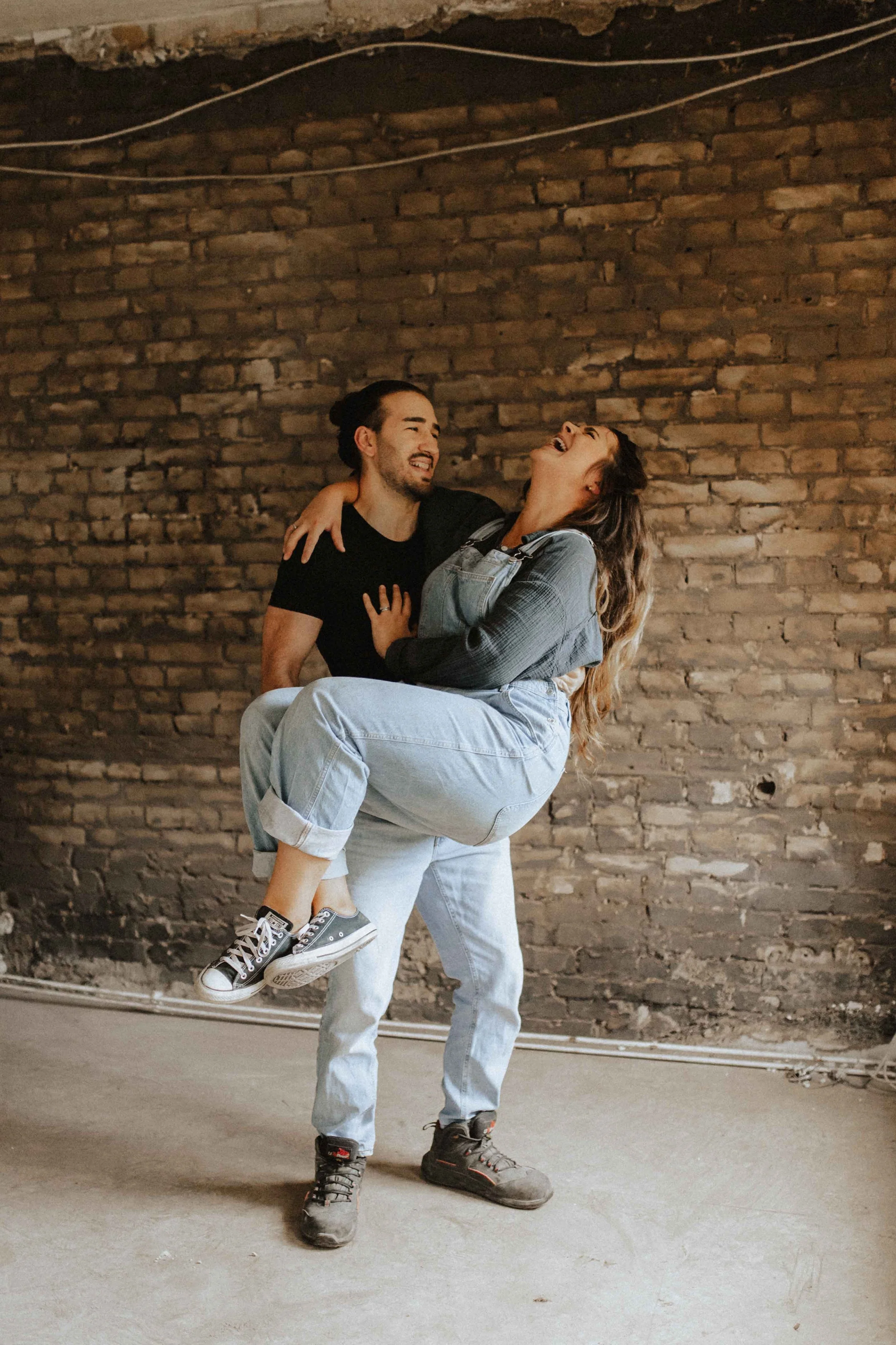 Paarshooting auf Baustelle – lachendes Paar vor Backsteinwand, der Mann trägt die Frau auf dem Arm, beide schauen sich fröhlich an, in Lüneburg.