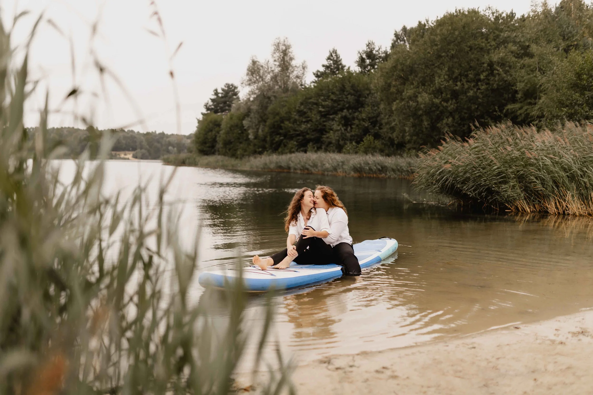 Zwei Frauen sitzen umarmt auf einem SUP-Board am See, eingerahmt von Schilf und Bäumen
