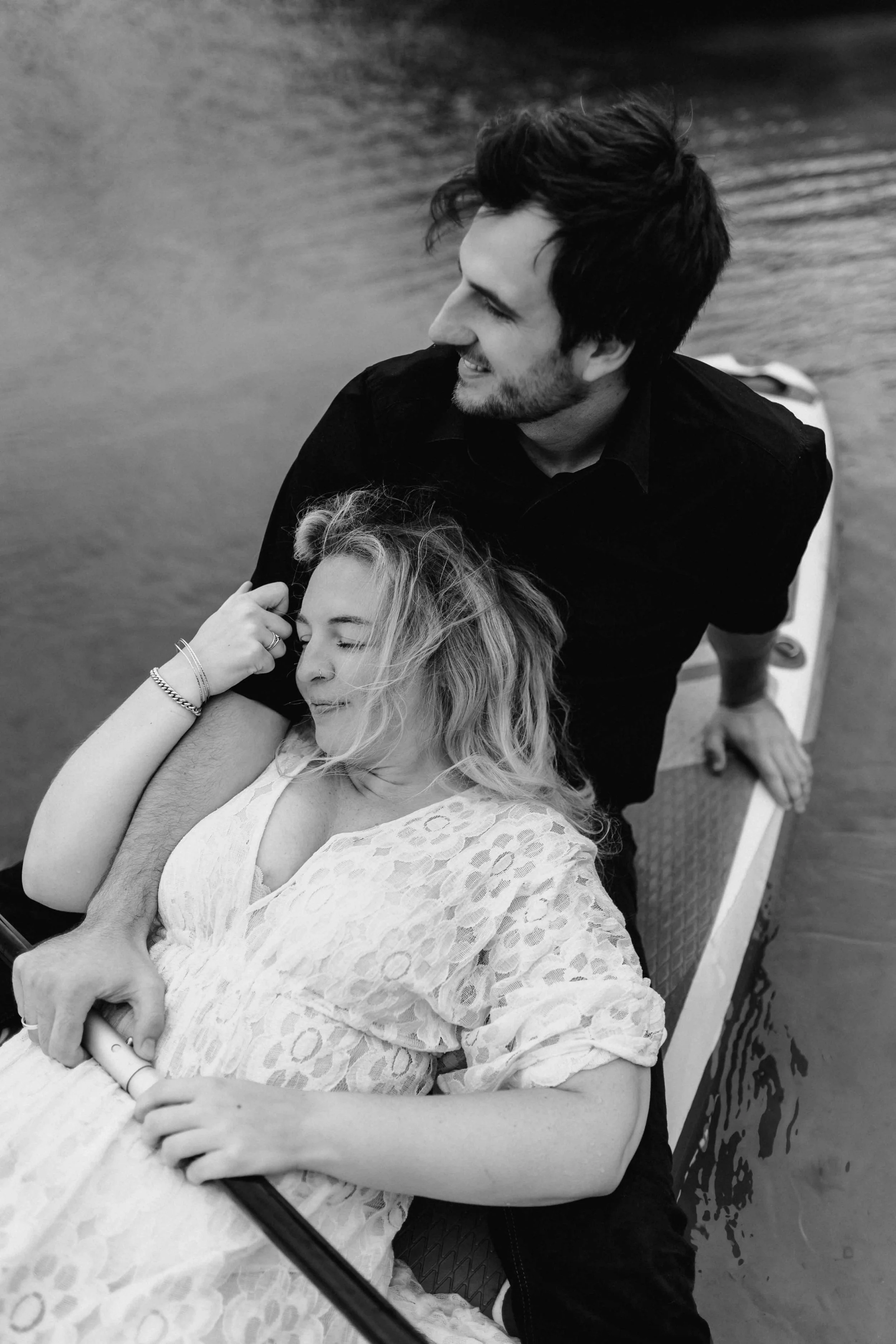 Paar sitzt auf einem SUP-Board am See, sie lehnt sich entspannt an seine Schulter, schwarz weiß Fotografie