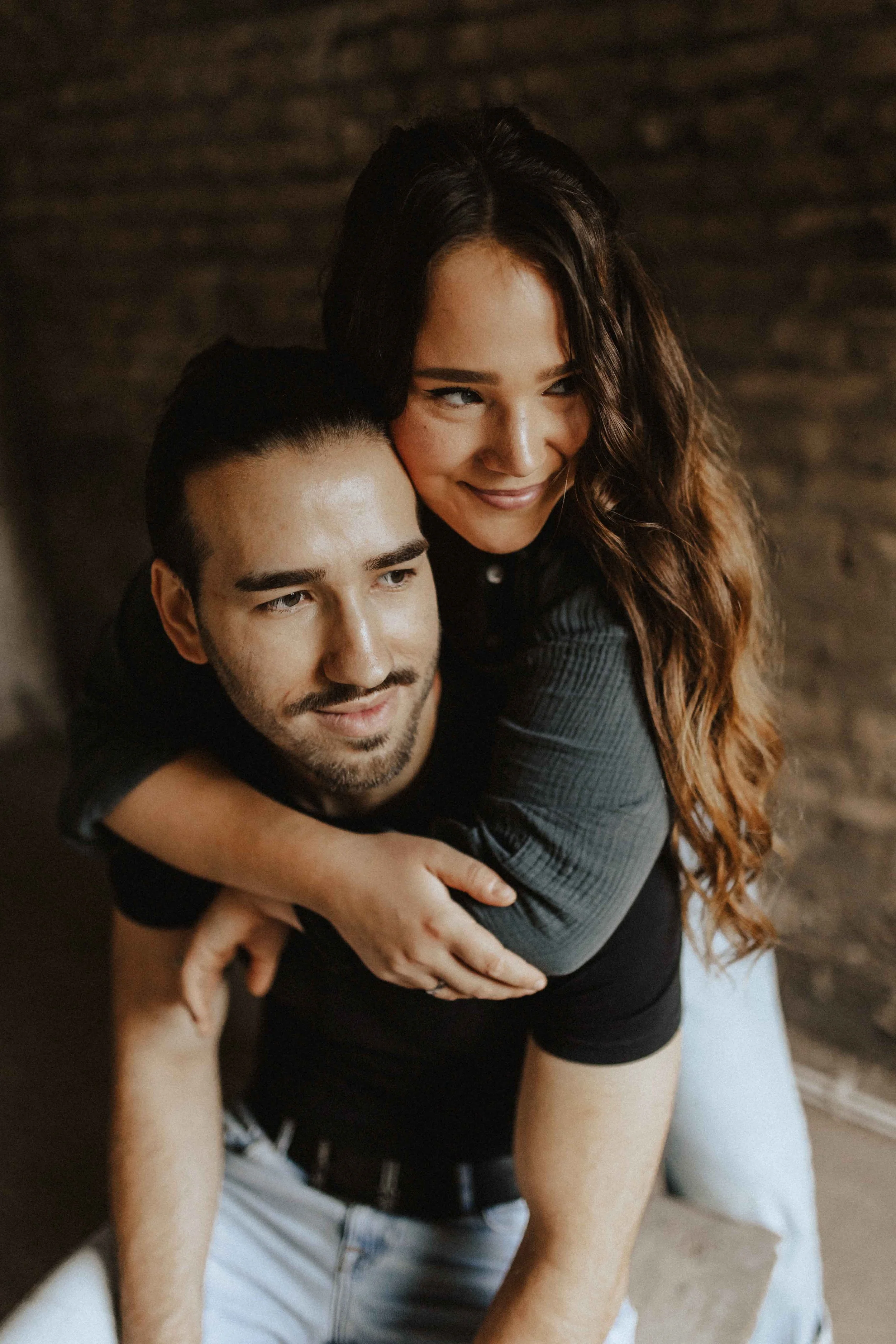 Paarshooting in Uelzen – Frau umarmt ihren Partner von hinten und lächelt verschmitzt, beide vor einer Backsteinwand.