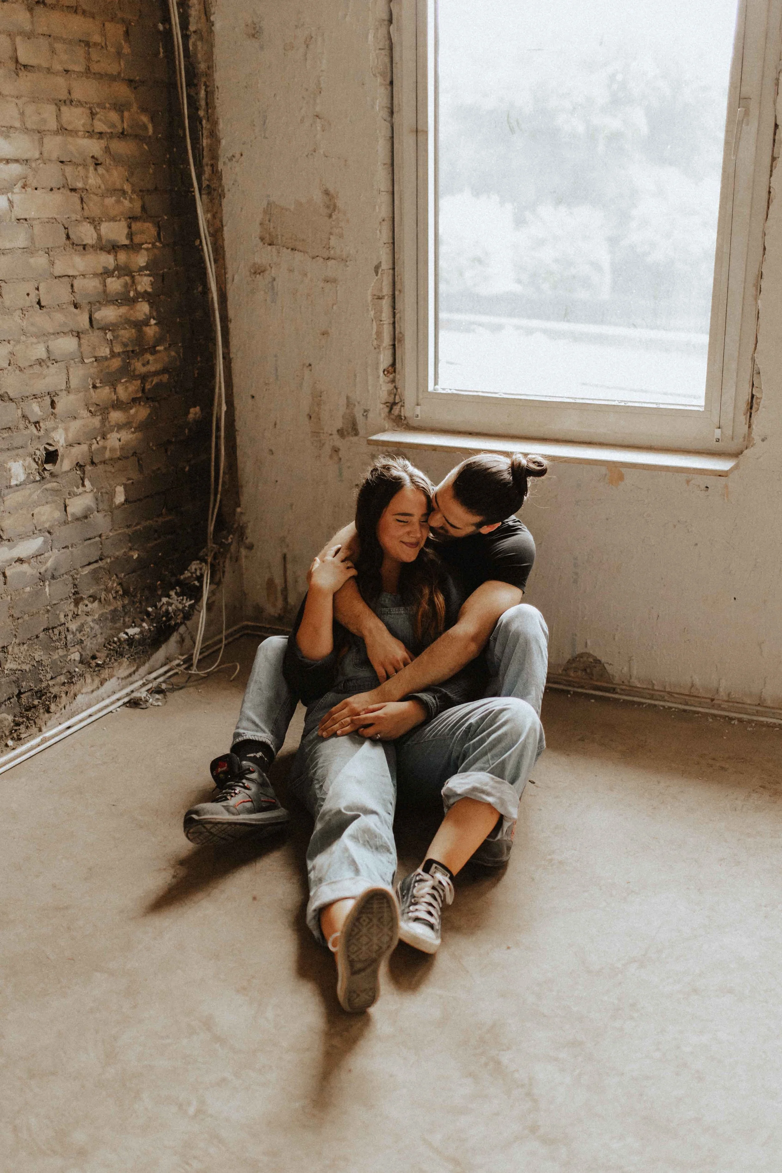 Paarshooting auf Baustelle – Paar sitzt eng umschlungen auf dem Boden vor einem Fenster in Lüneburg und genießt den Moment.