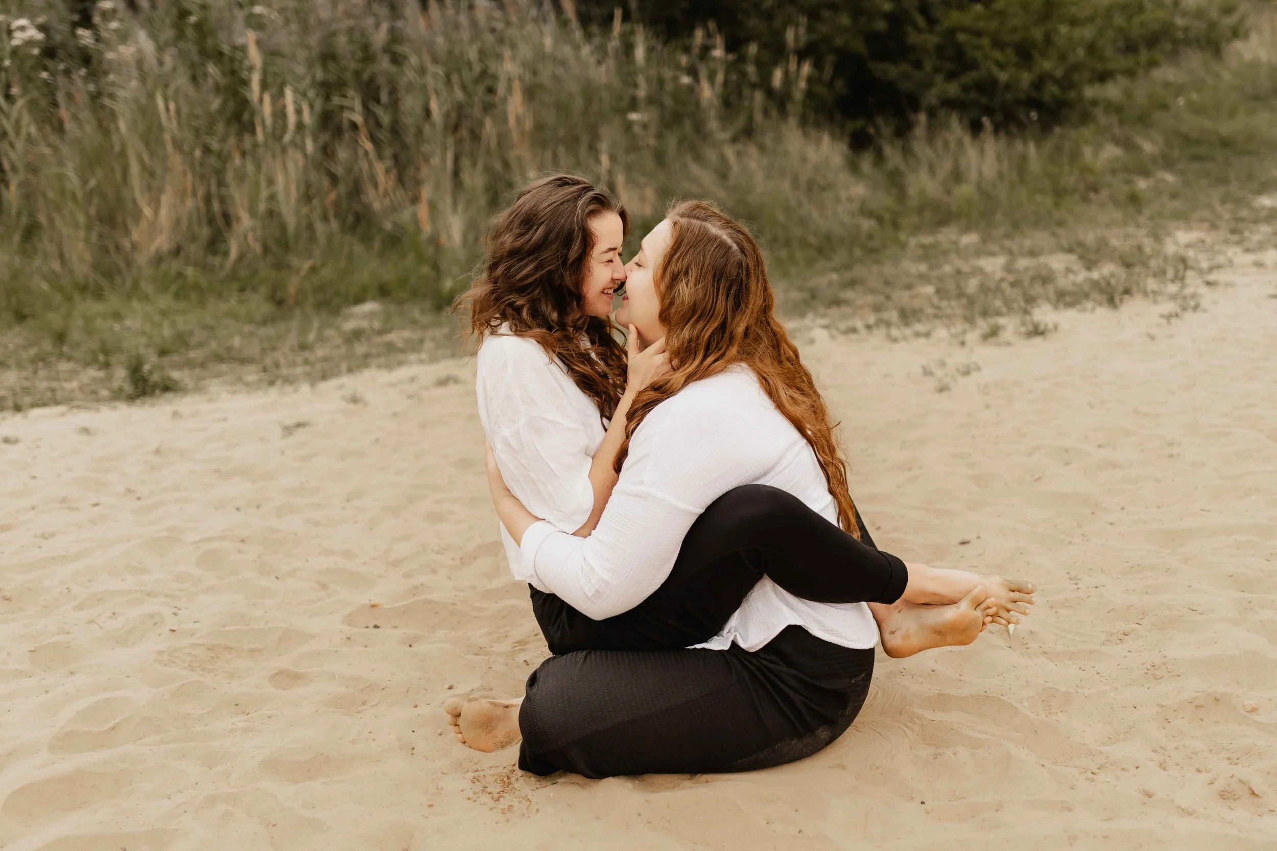 Verliebtes Frauenpaar sitzt eng umschlungen im Sand und küsst sich