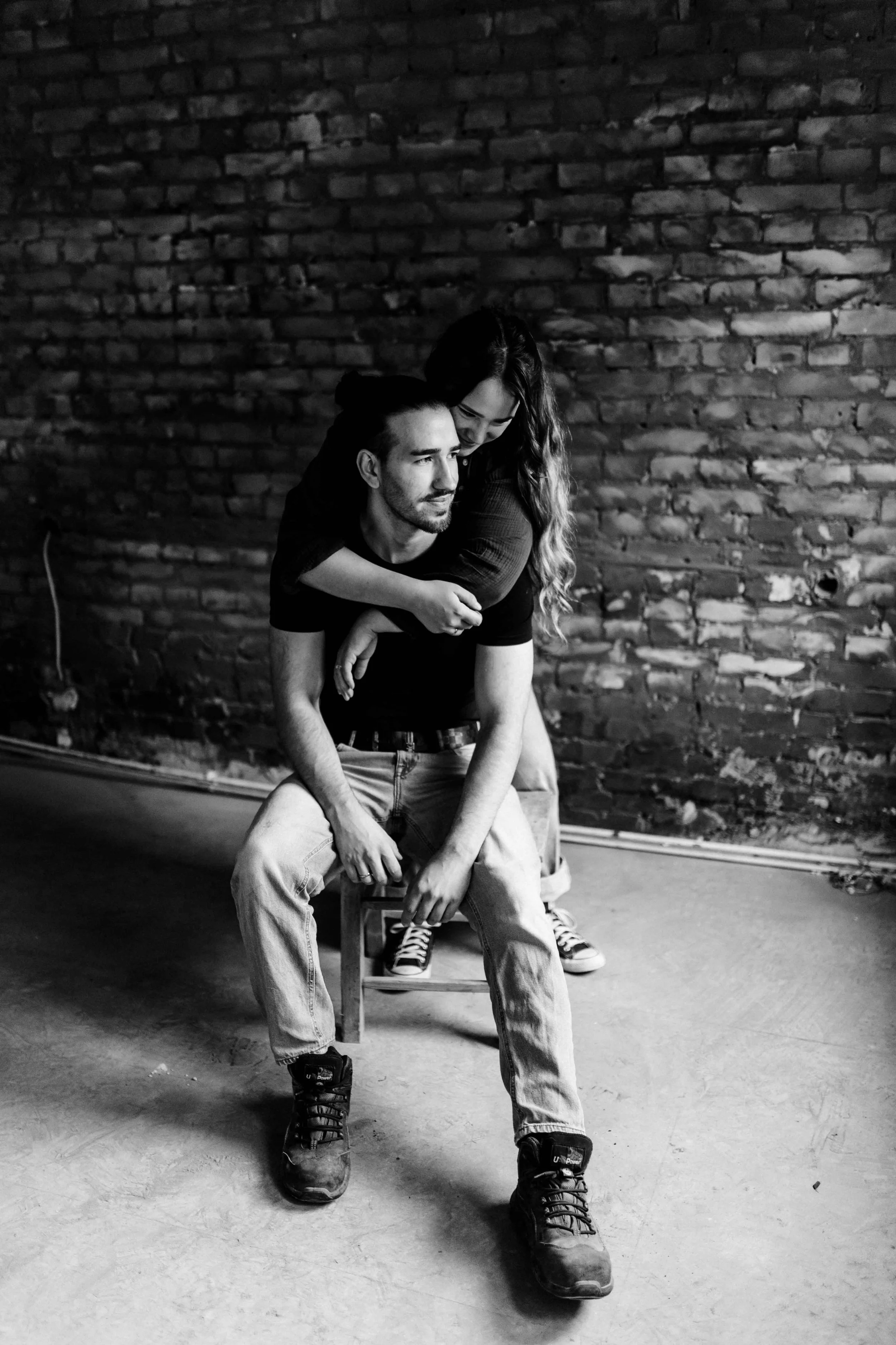 Schwarz-weiß Paarshooting in Lüneburg – Frau umarmt ihren sitzenden Partner von hinten vor einer rustikalen Backsteinwand.