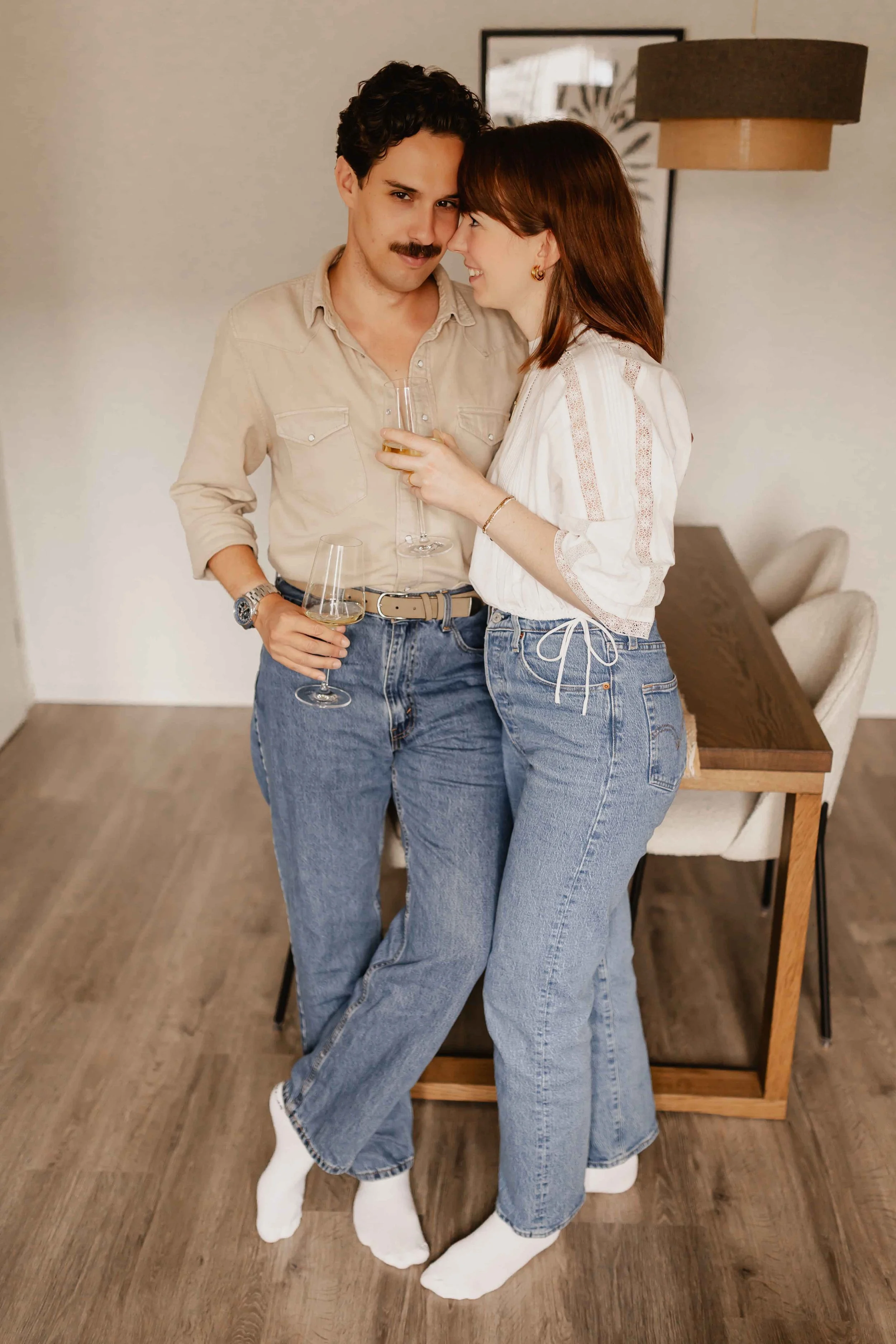 Paar steht bei einer Homestory in Lüneburg eng beieinander im Wohnzimmer, er lehnt lässig am Tisch, sie schaut ihn verliebt an