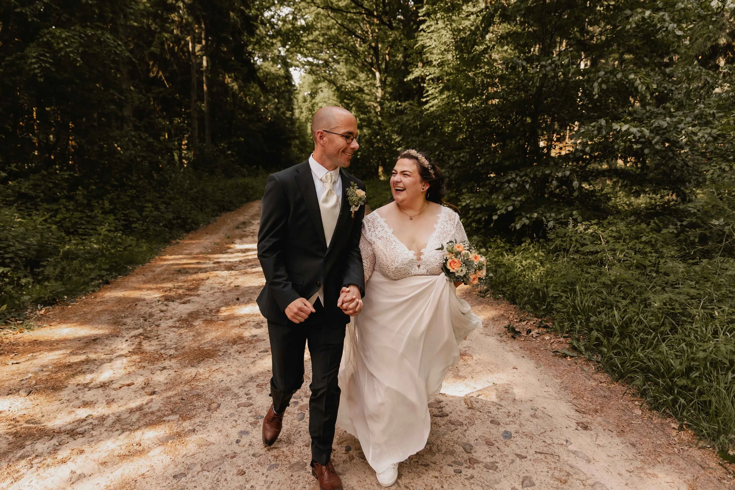 Hochzeitspaar spaziert lachend Hand in Hand durch den Wald