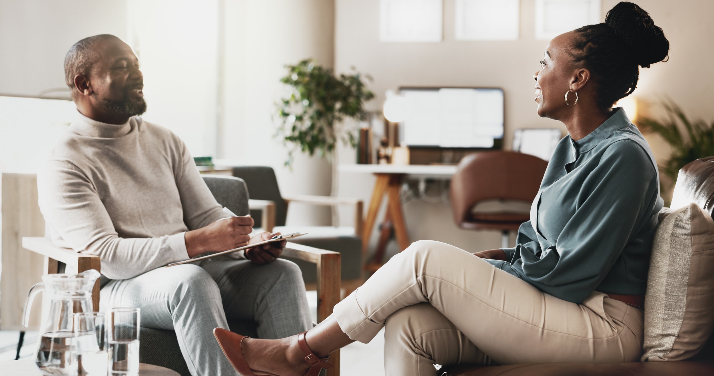 A woman and a man are having a conversation in a sunlit living room with large plants, a glass coffee table, and a window in the background.