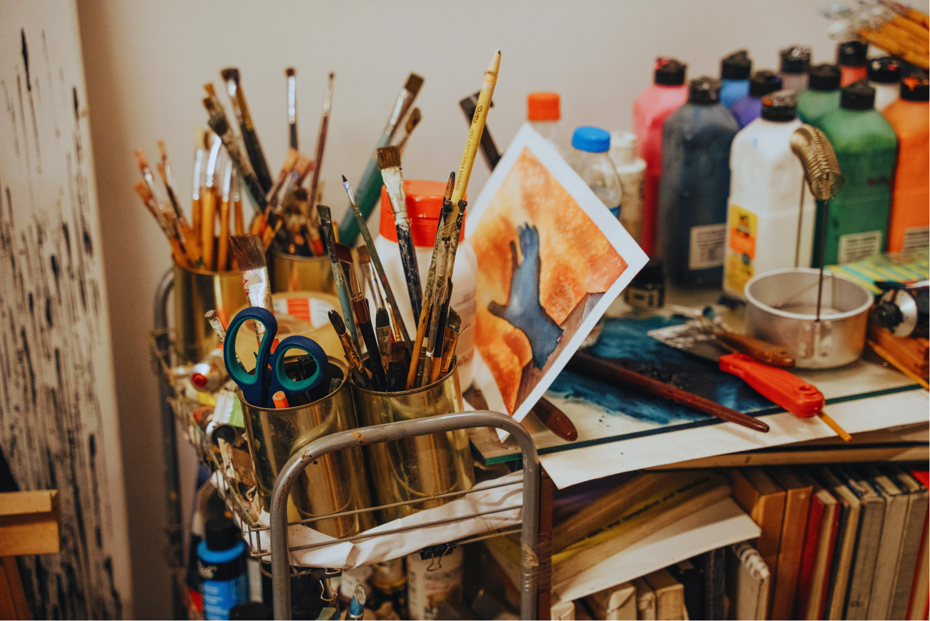 An artist's workspace featuring paintbrushes in metal containers, watercolor paintings, bottles of paint, and various art supplies on a cluttered table.