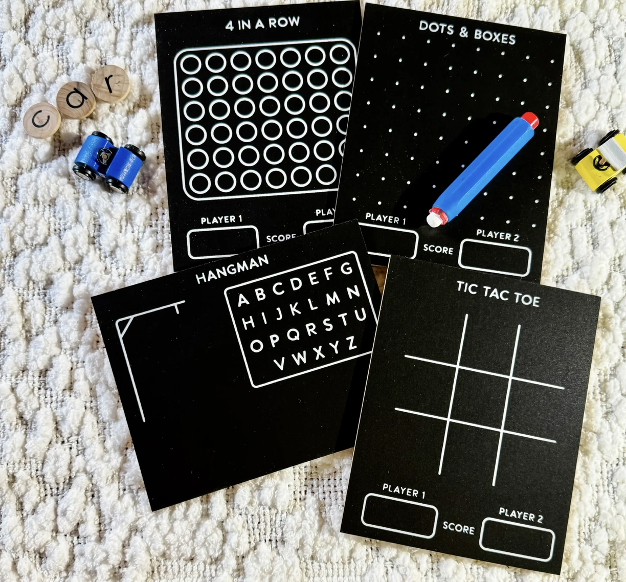 Black game boards laid out with titles "4 In A Row," "Dots & Boxes," "Hangman," and "Tic Tac Toe" on a textured surface. Wooden letter discs spelling "car" and small toy cars are nearby.