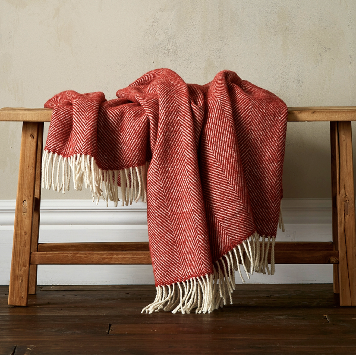 Red herringbone-patterned throw blanket draped over a wooden bench with white fringes.
