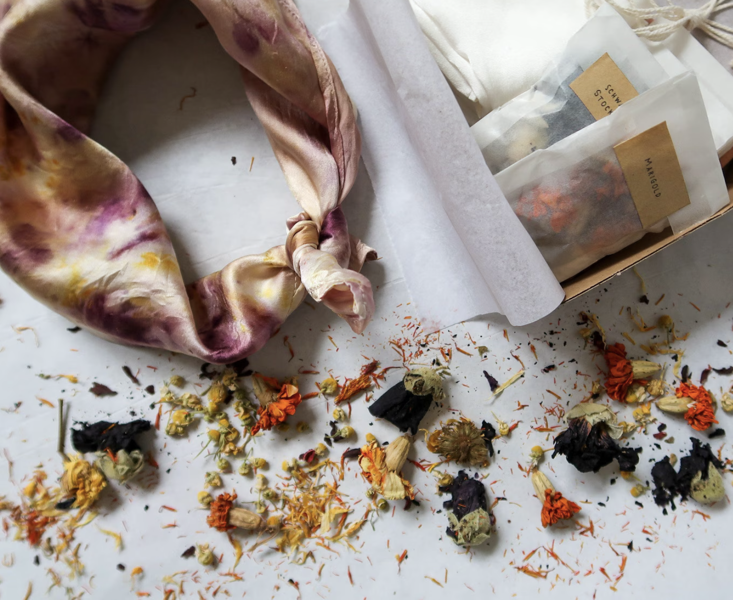 A tie-dye silk scarf, dried flowers, and labeled seed packets placed on a white surface.