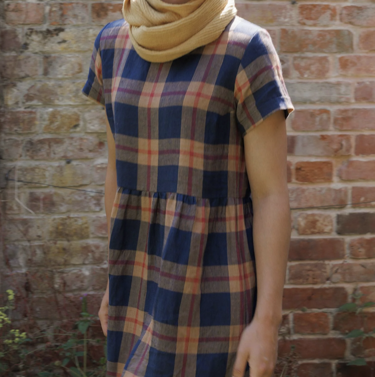 Person wearing a plaid dress and scarf in front of a brick wall.