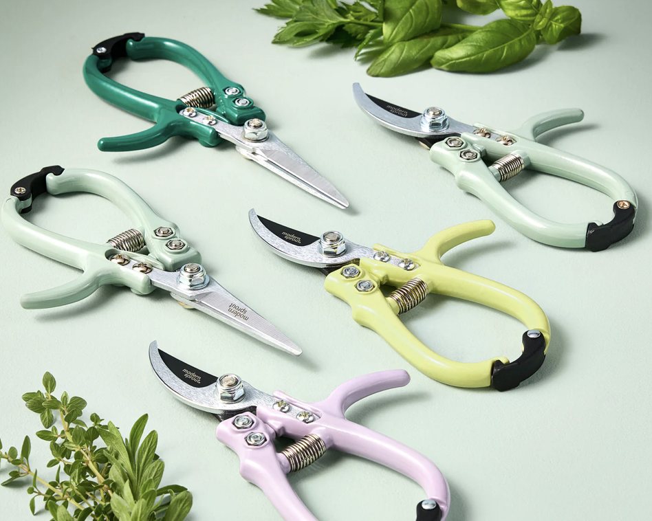Four colorful garden pruners on a light surface with fresh herbs in the background.