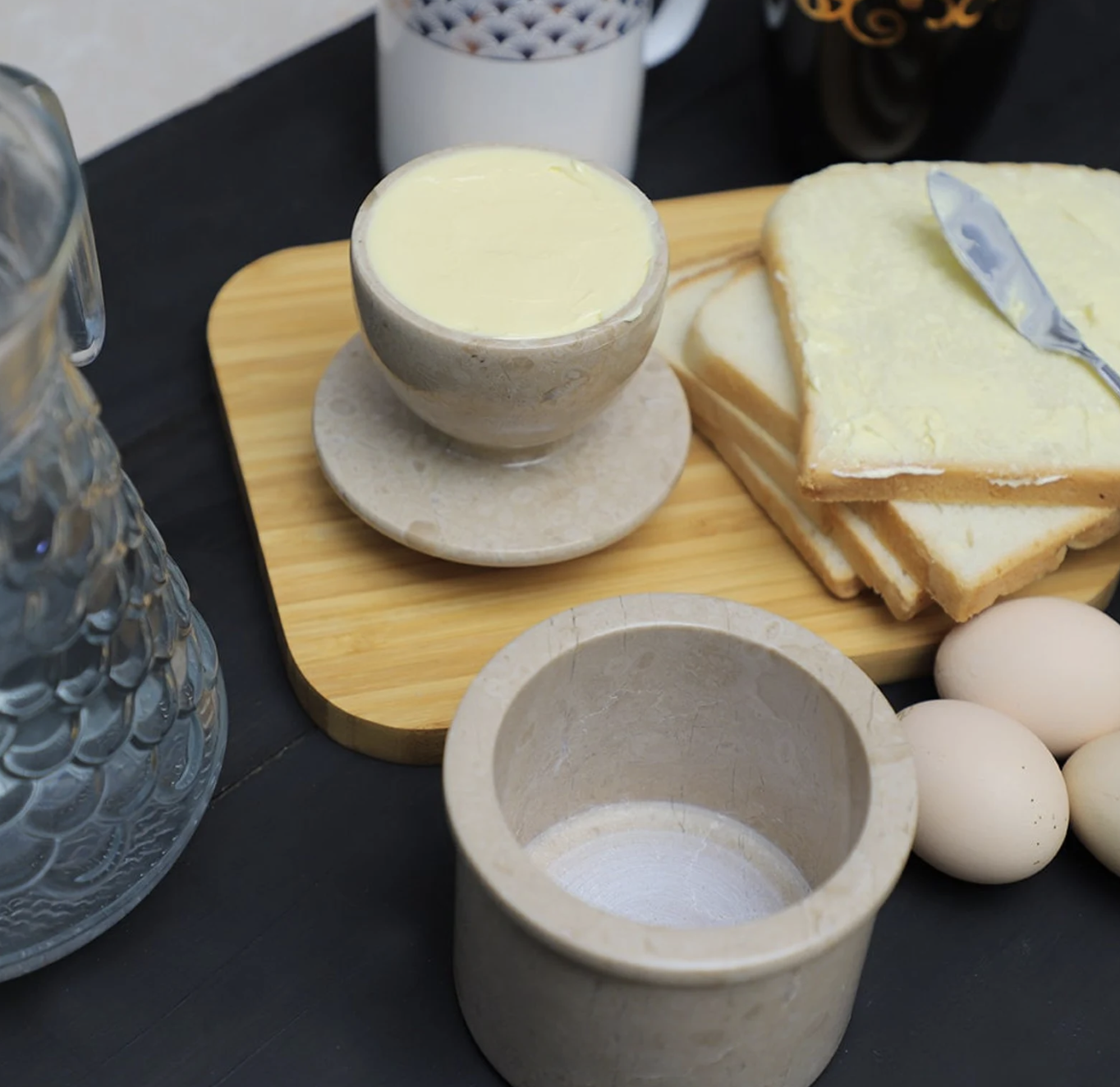 Butter in a cup, sliced bread with butter on a wooden board, eggs, and glass jug on a dark surface.