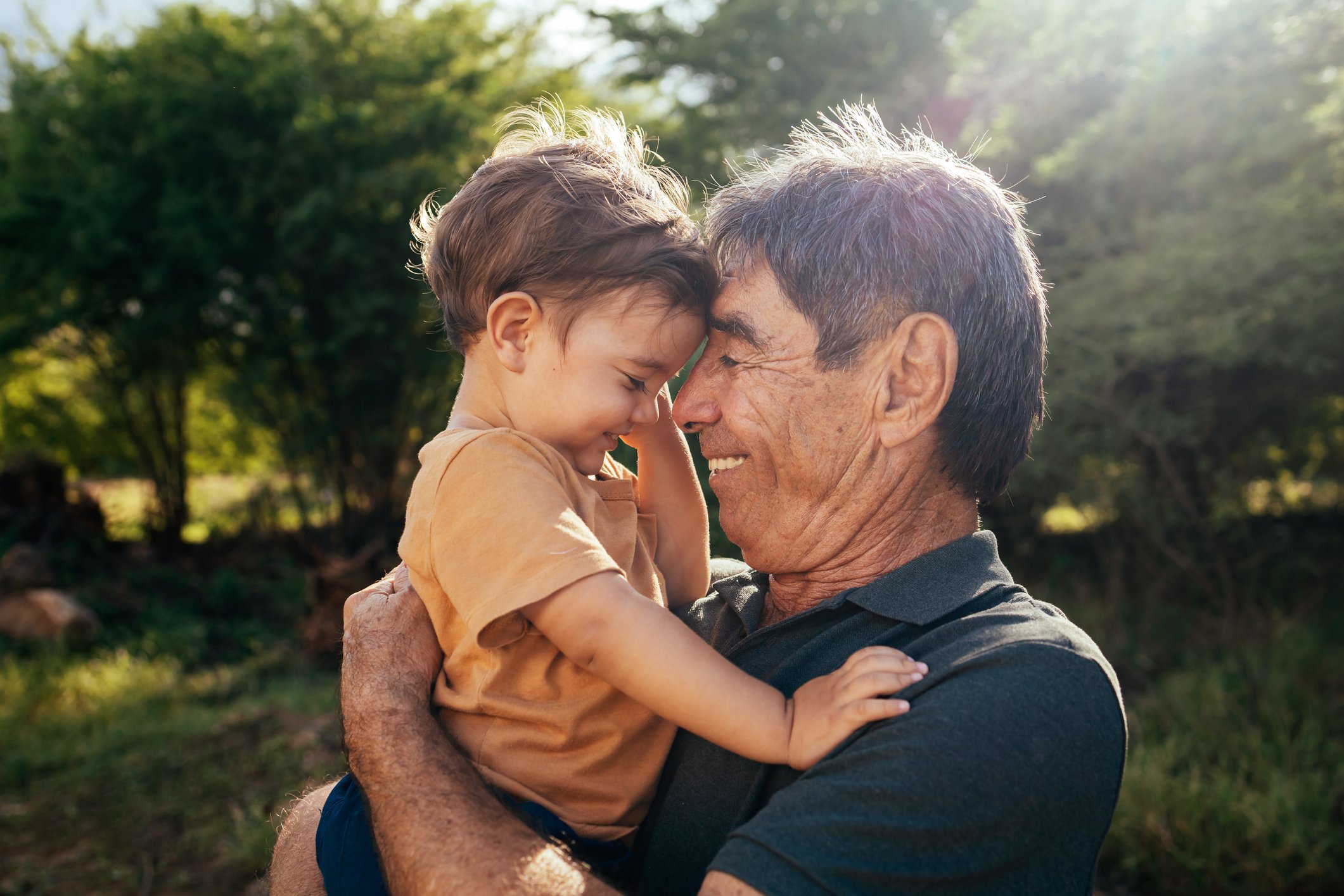 An elderly man holding a young boy close, with their foreheads touching in a loving embrace outdoors, surrounded by trees.