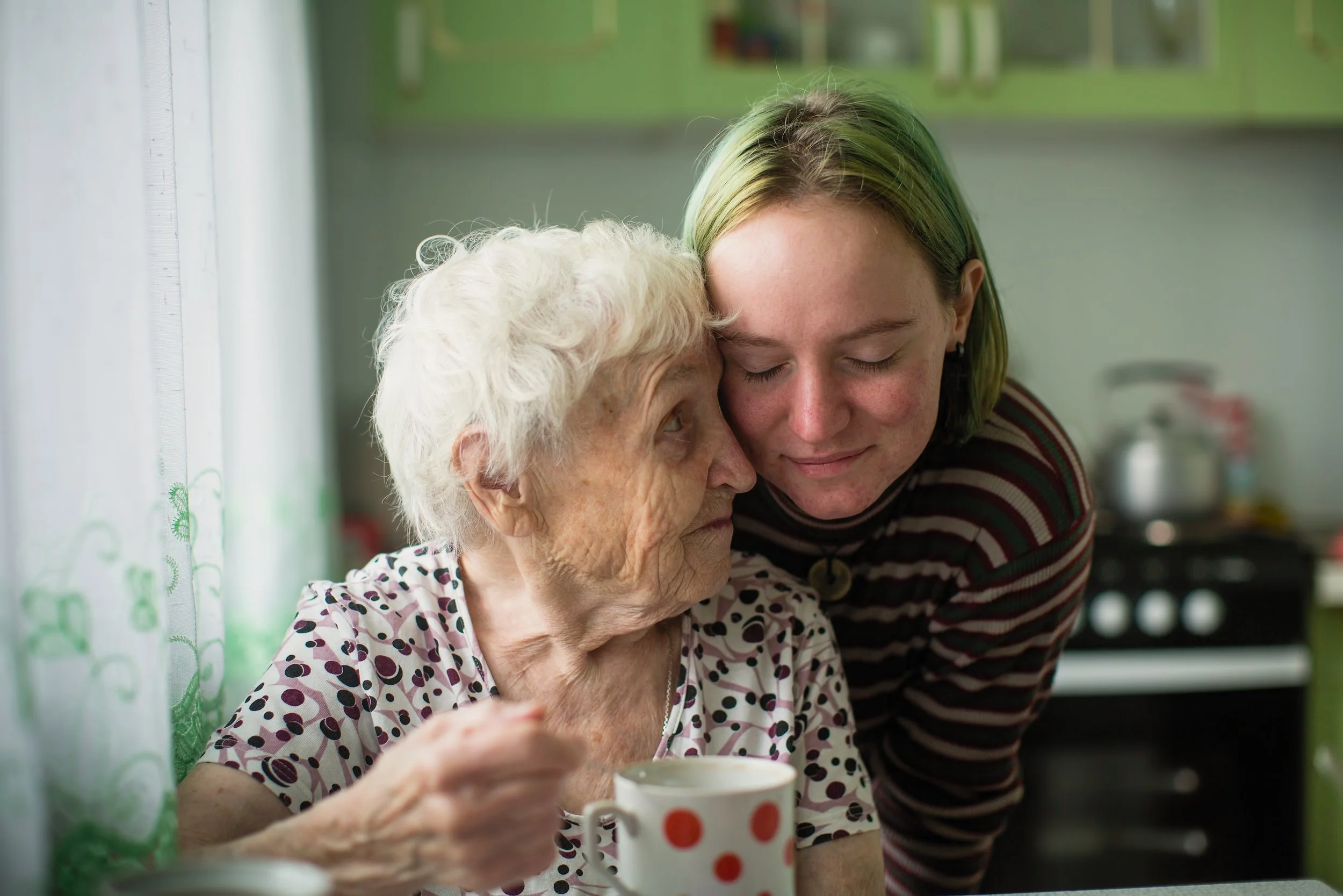 An elderly woman with short, curly white hair and a young woman with shoulder-length blonde hair in a striped shirt show affection by resting their foreheads together while sitting by a window with green patterned curtains. The elderly woman holds a mug with red polka dots.