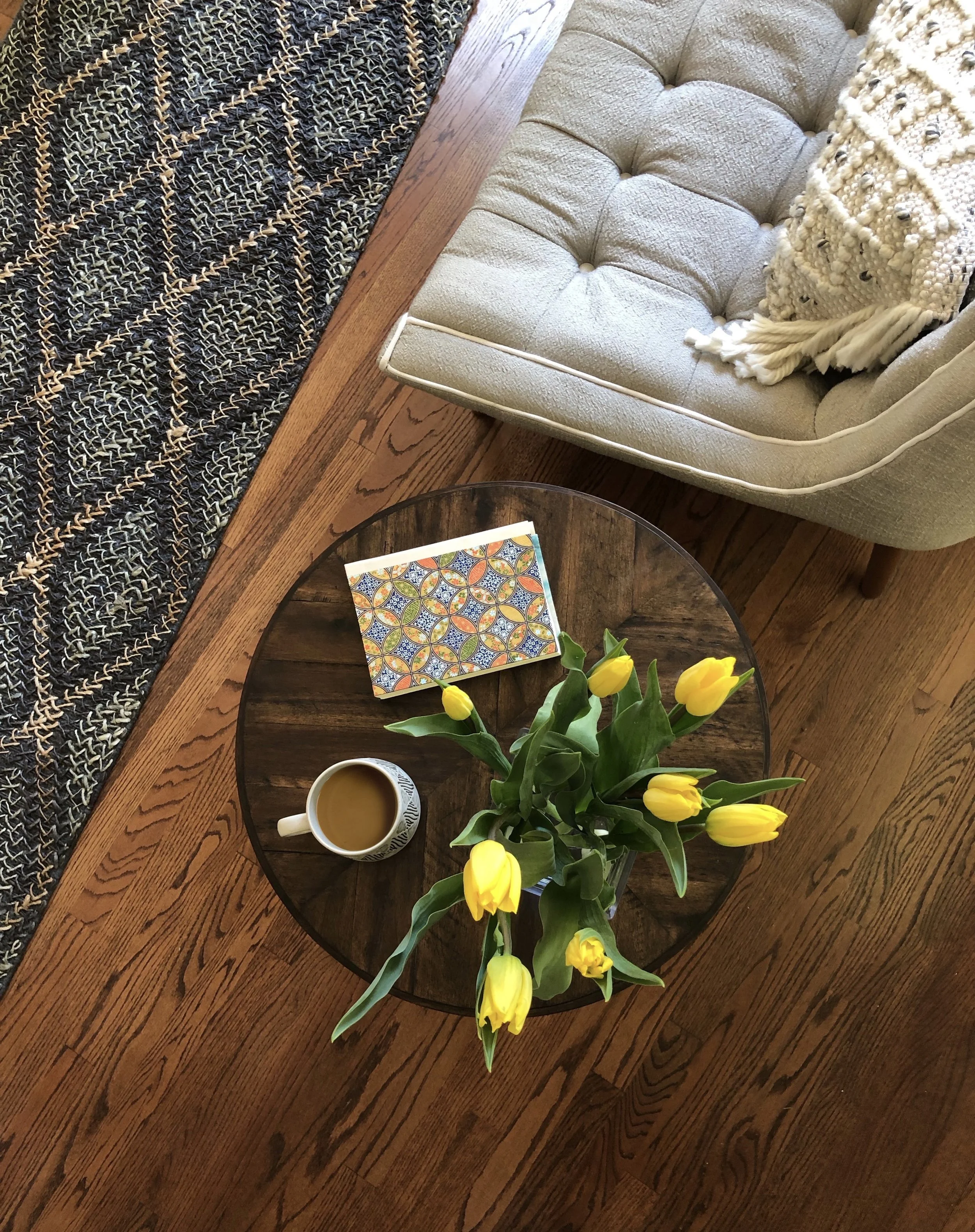 A round wooden table with a vase of yellow tulips, a cup of coffee, and a colorful patterned notebook. A beige tufted armchair and a decorative pillow are visible near the table. The setting appears to be a cozy living room or sitting area with hardwood flooring.