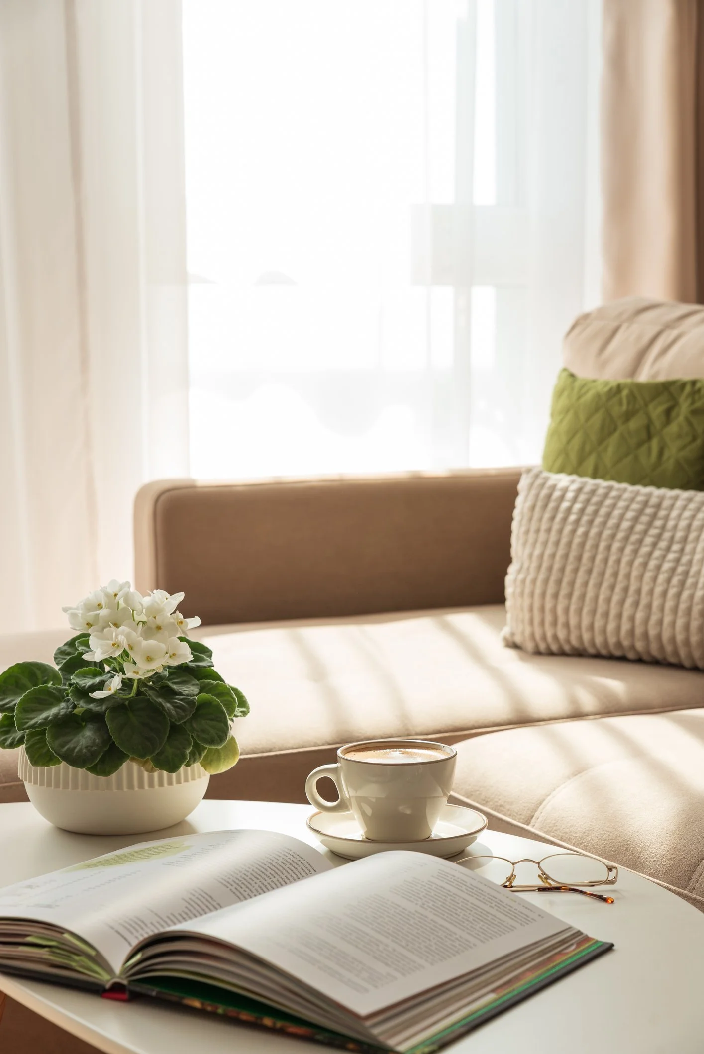 A cozy indoor scene featuring an open book, a white cup of coffee or tea on a saucer, a pair of reading glasses, and a potted plant with white flowers on a white table. In the background, there is a beige sofa with green and striped pillows, a window with sheer curtains, and a softly lit room.