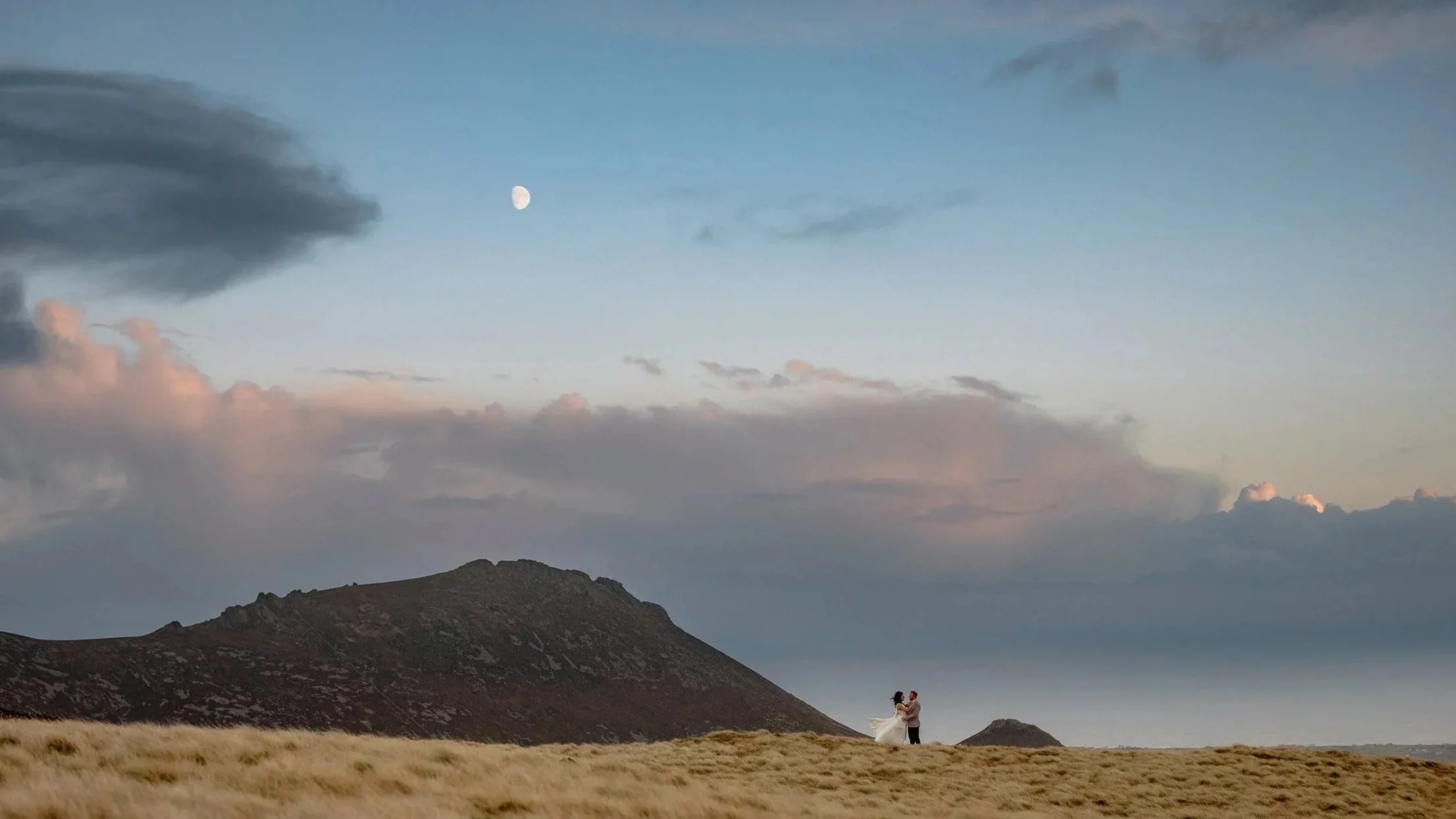 A couple in wedding attire standing in a grassy field near a mountain with a cloudy sky and the moon overhead.
