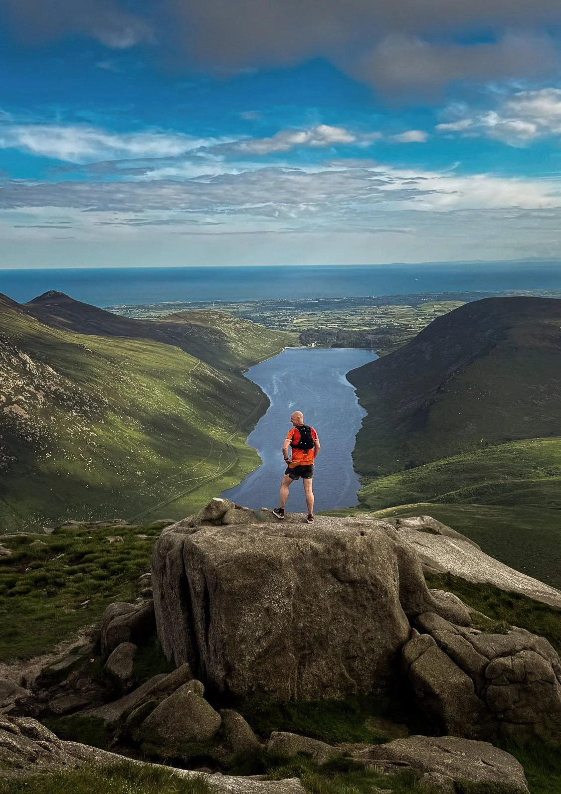 A person standing on a large rock overlooking a scenic valley with a river and rolling hills, ocean in the background, under a partly cloudy sky.