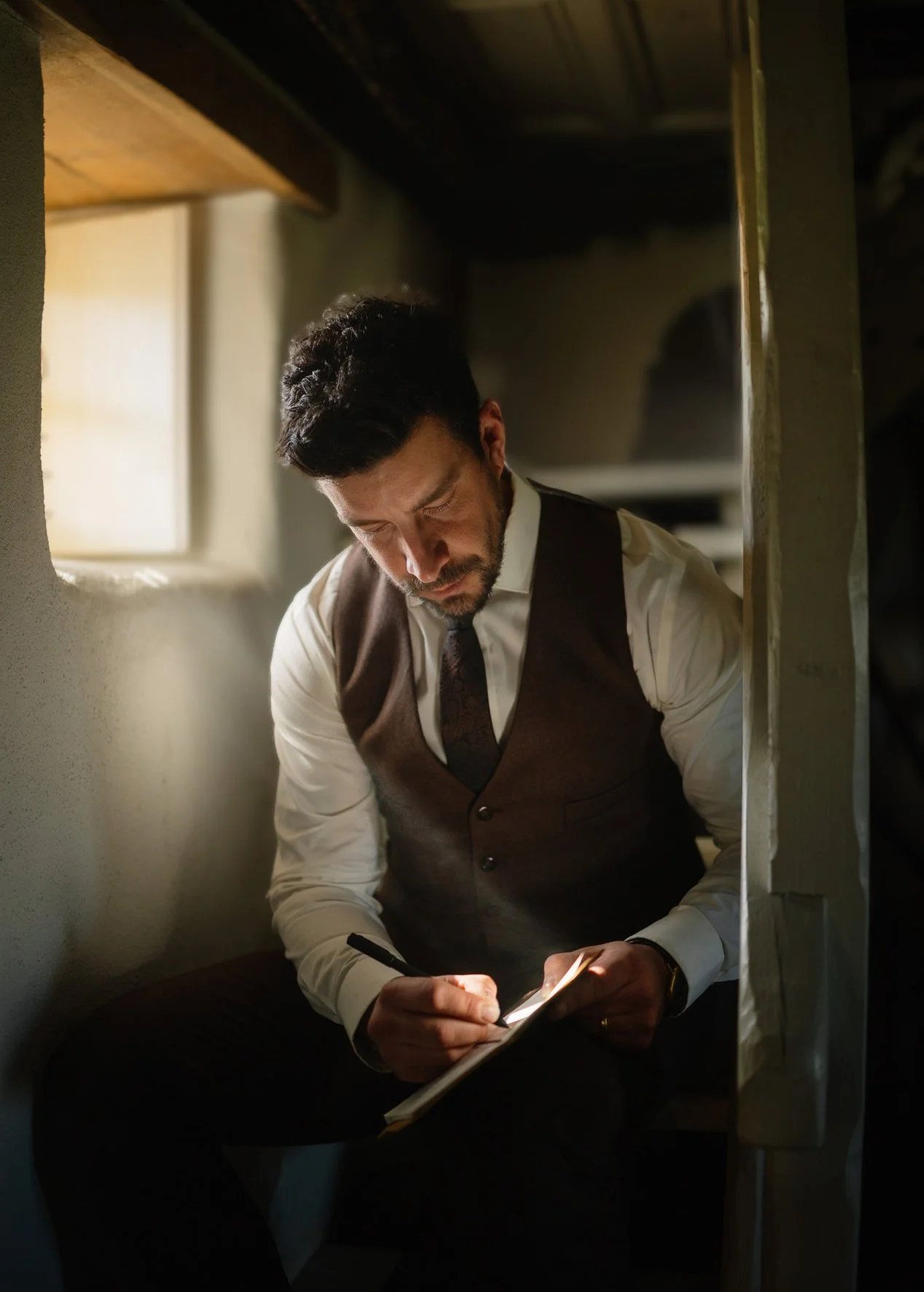 A man dressed in a white shirt, dark vest, and tie is writing on a notepad inside a small, dimly lit room or booth, with light coming from a device he's holding.