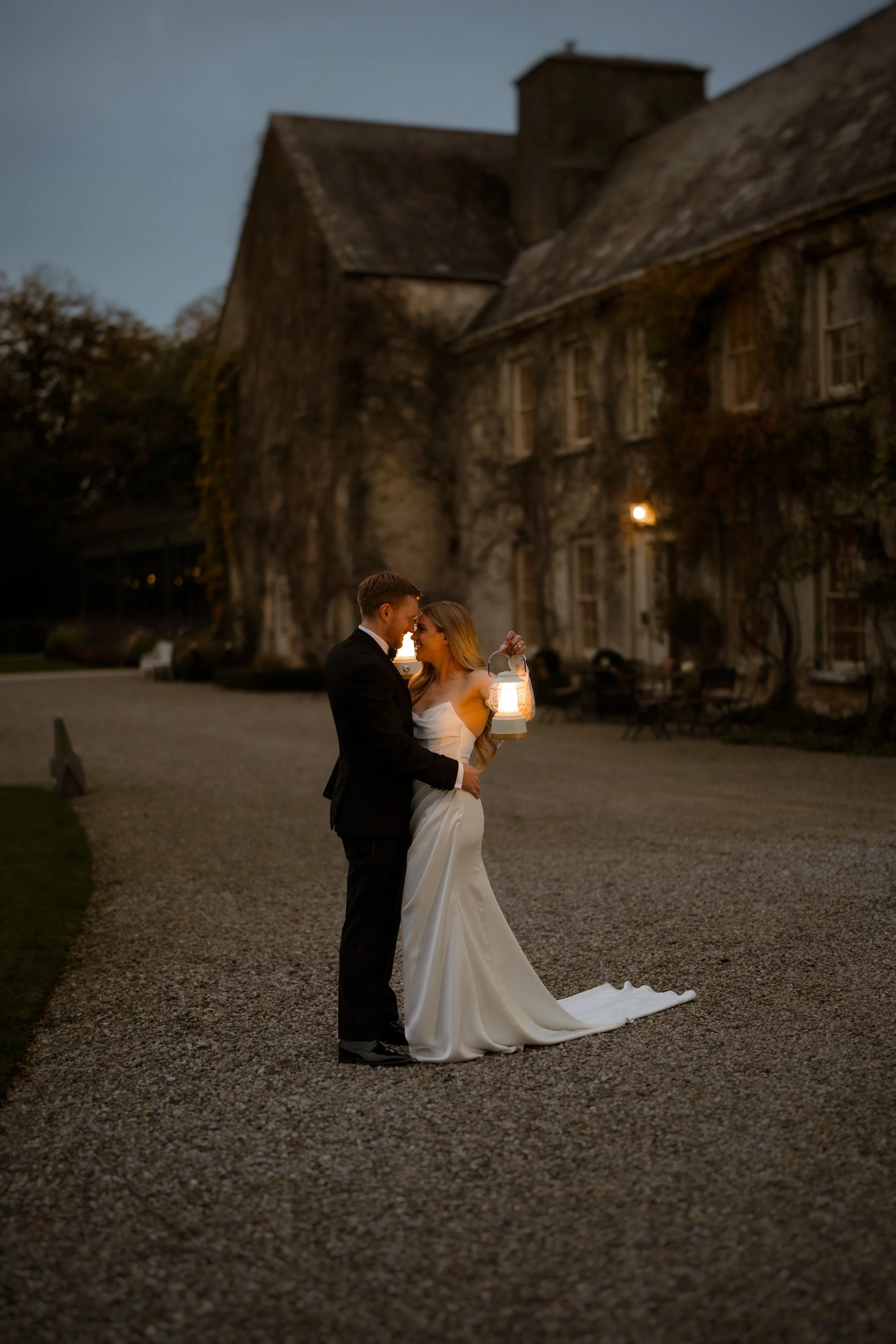 A bride and groom at night, standing on a gravel path outside a large, old brick building with ivy-covered walls, holding a lantern.