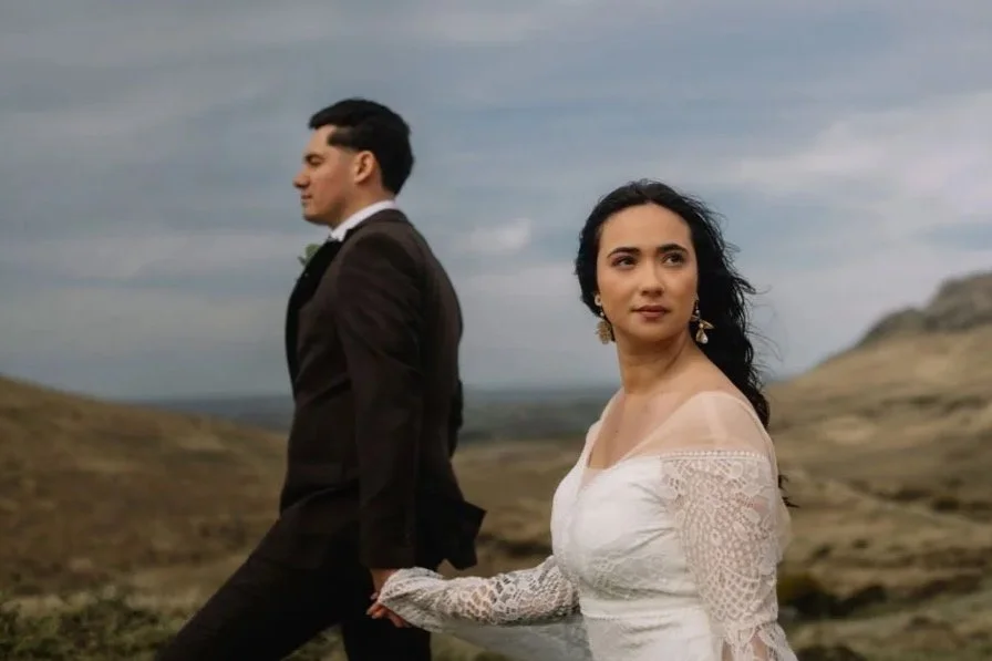 A man and a woman stand in a barren, hilly landscape. The woman is in the foreground, wearing a white lace dress, looking to the right with a contemplative expression. The man is in the background, wearing a black suit, facing away with his eyes clos