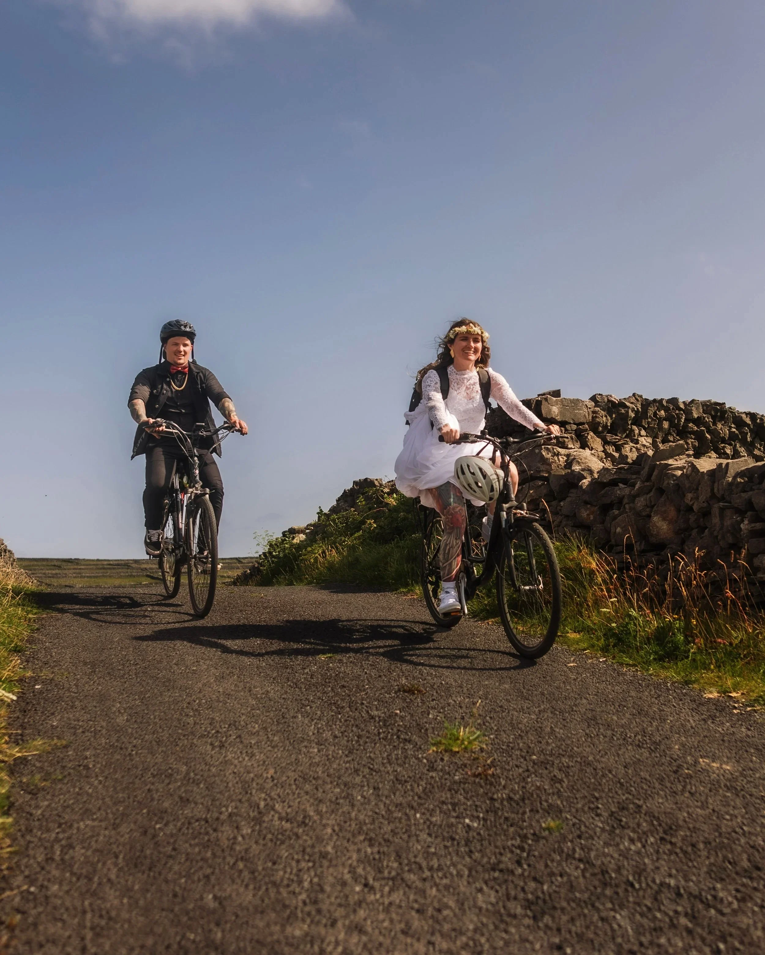 Elope couple riding bicycles on a rural path, one wearing a white dress and floral headband, the other in dark casual clothing, with a clear sky and stone wall in the background.