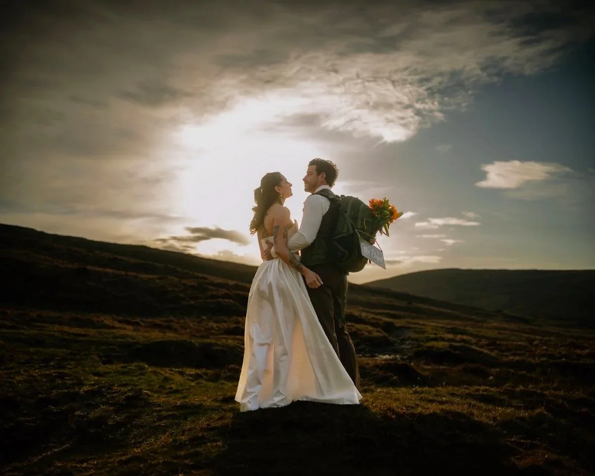 A couple in wedding attire standing close together outdoors at sunset, with a hilly landscape behind them.