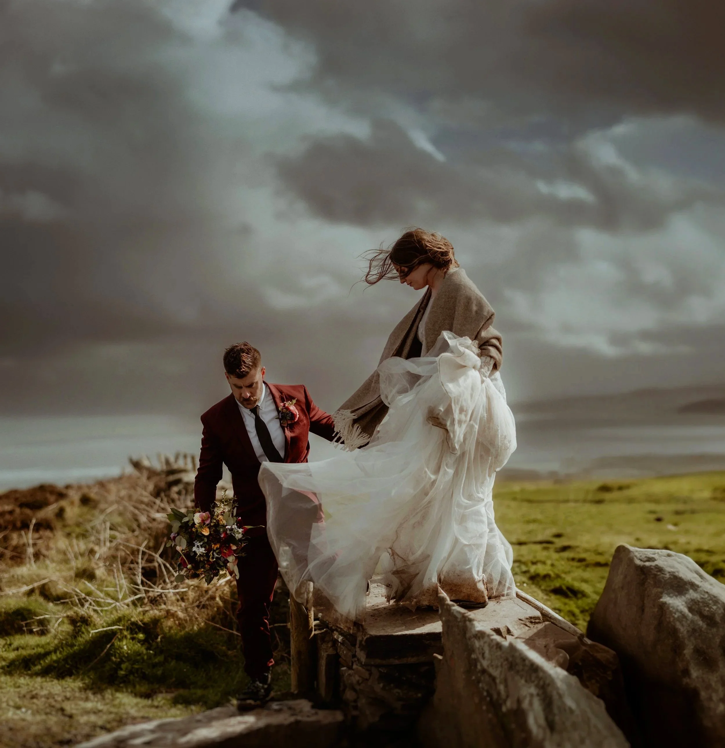Couple dressed in wedding attire outdoors on a rocky terrain. The woman is wearing a flowing white wedding dresst, standing on a stone structure, while the man, in a dark red suit, holds a bouquet of flowers and helps her balance at Cliffs of Moher