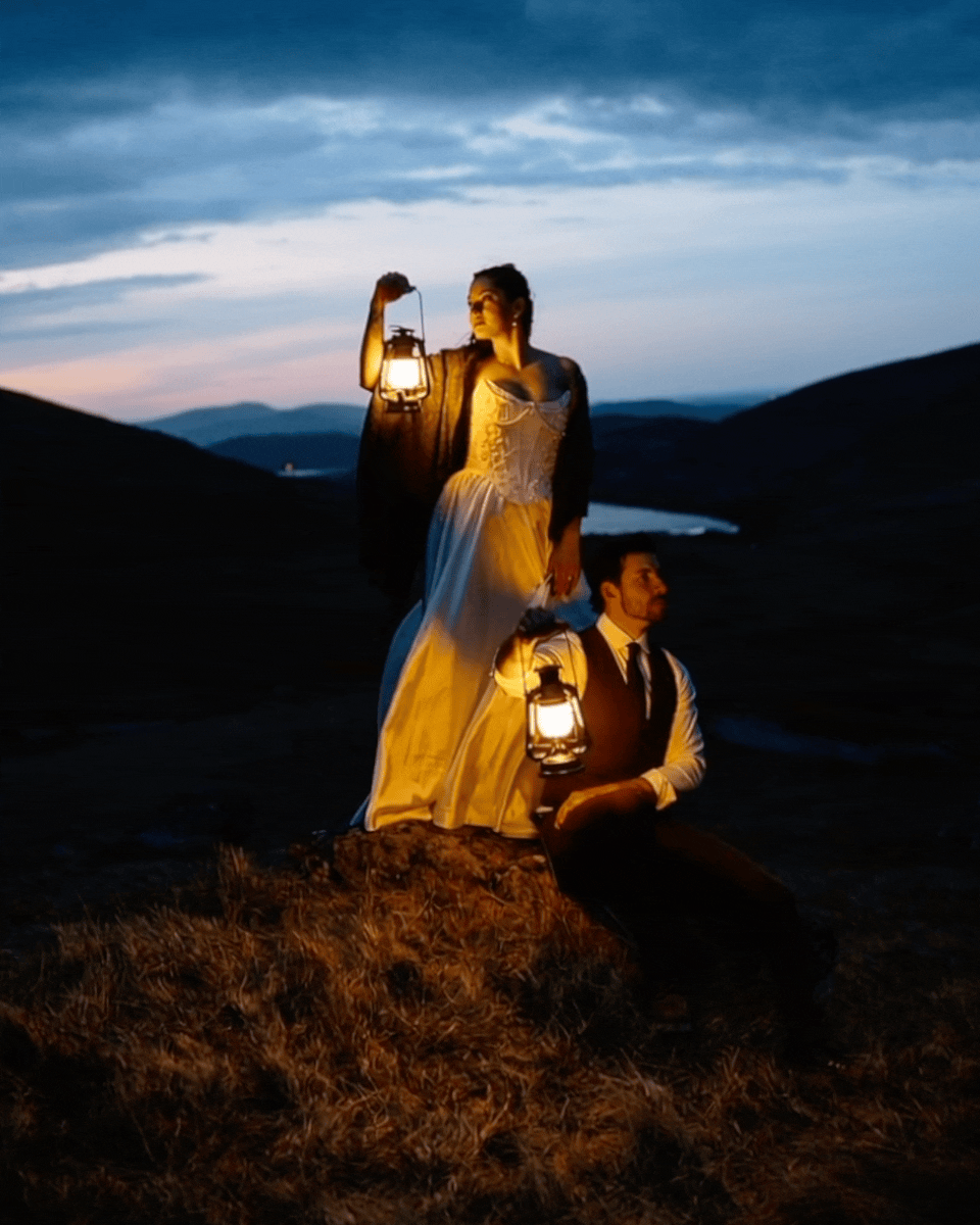 A woman and a man dressed in vintage clothing holding lanterns during dusk in a hilly landscape with a river in the background.