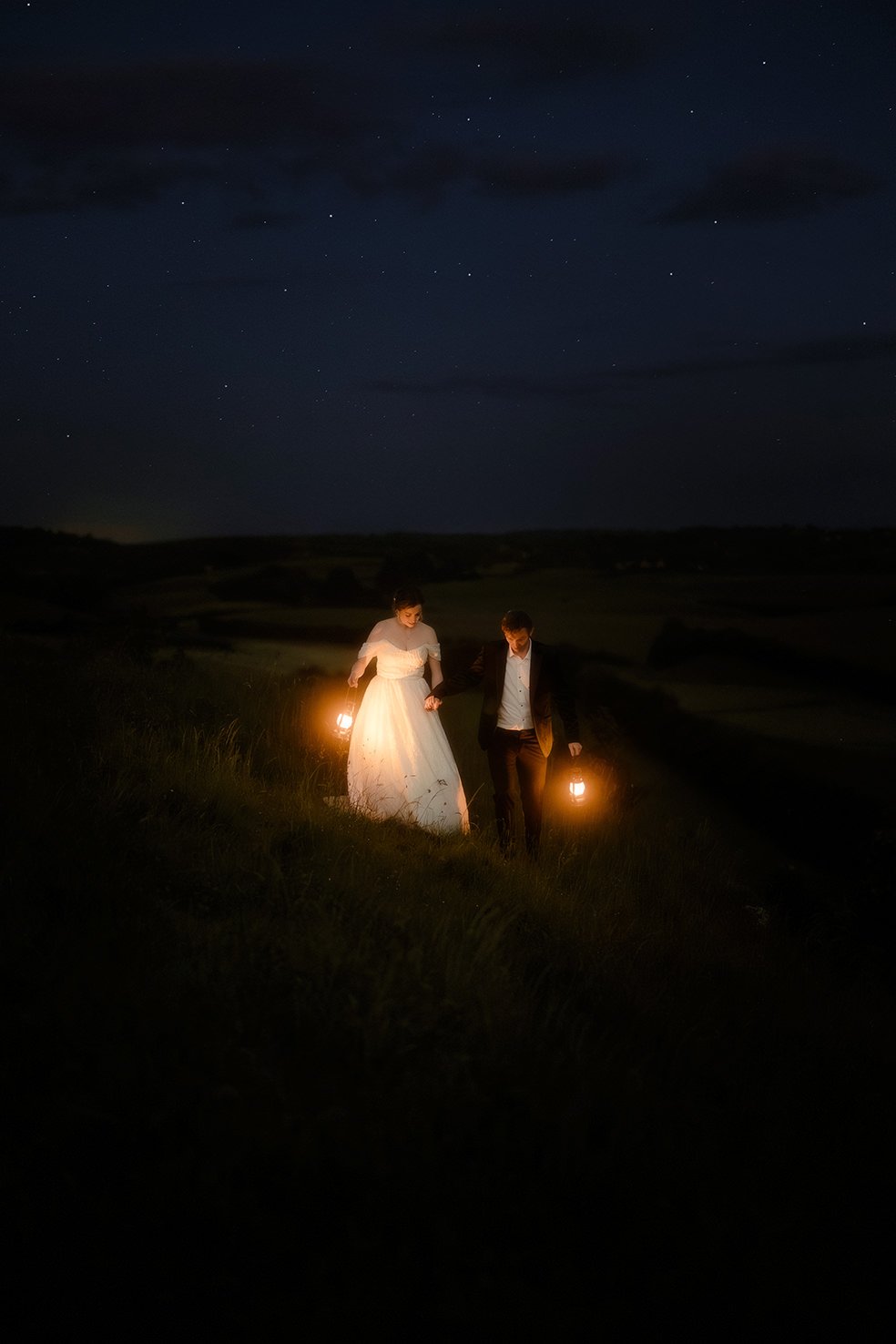 A couple, a woman in a white dress and a man in a suit, walking outdoors at night on a grassy hill, carrying lanterns, under a starry sky.