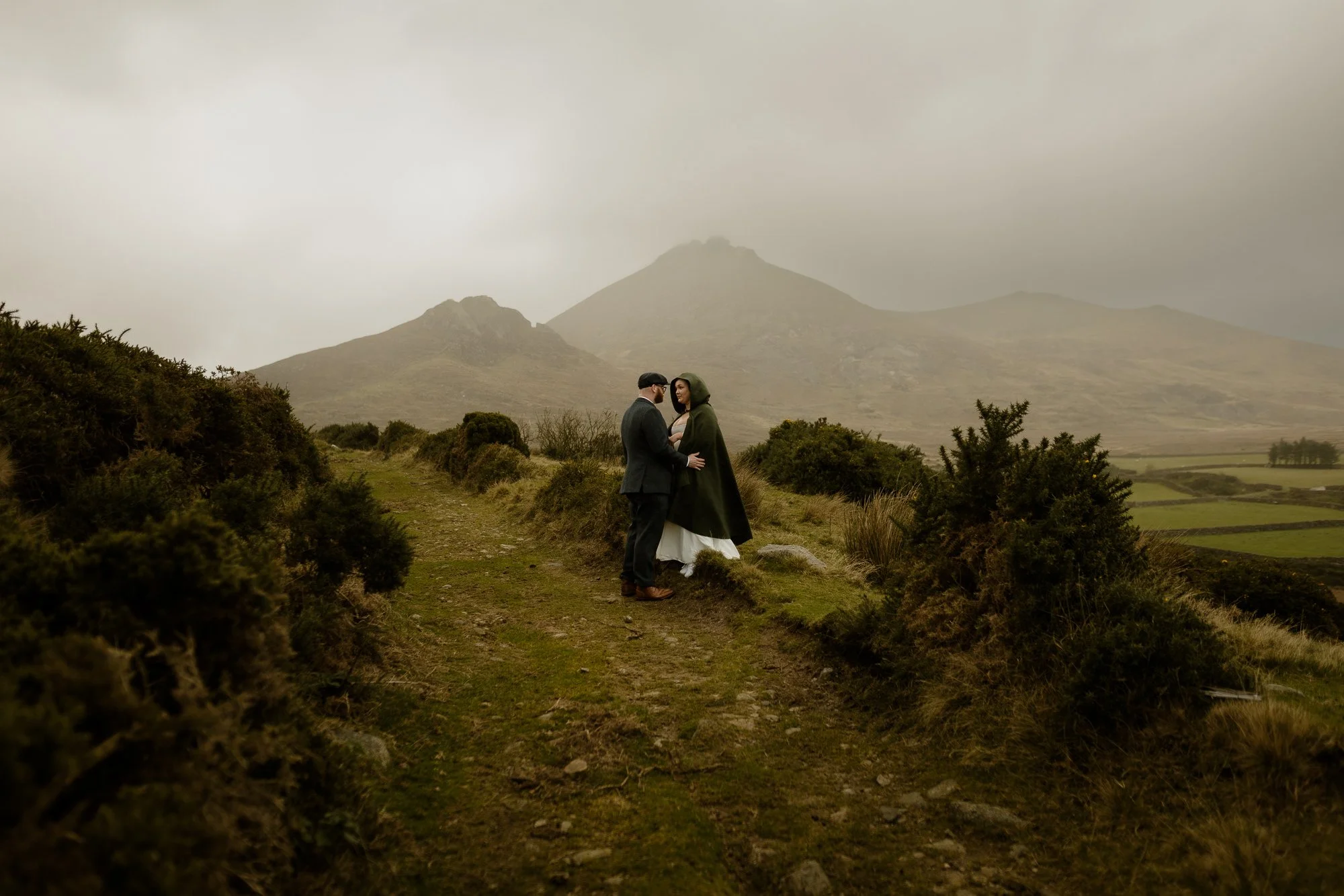 A couple dressed in vintage clothing standing on a dirt path in a mountainous landscape with cloudy weather.