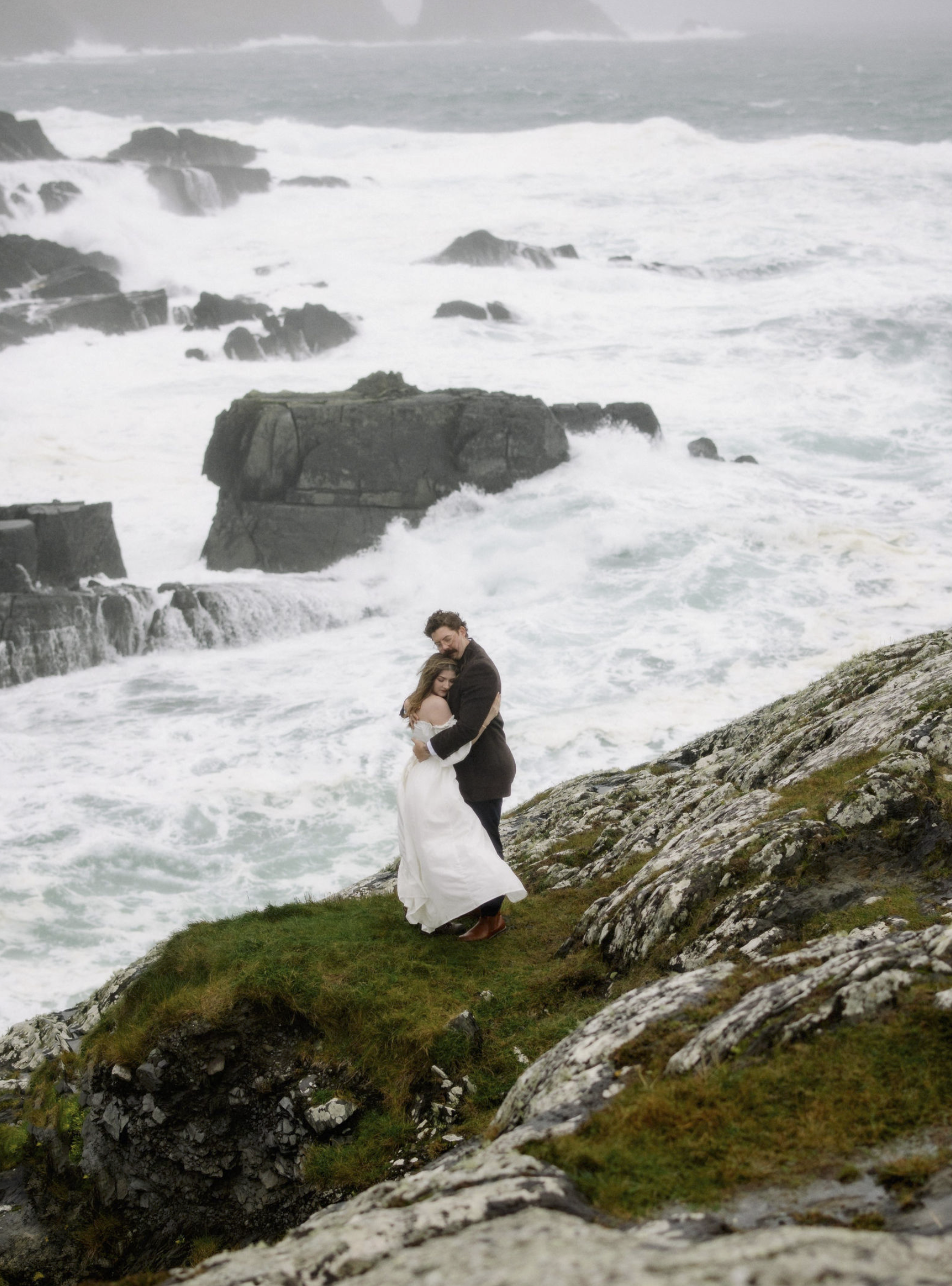A couple dressed in wedding attire embracing on a rocky coastline with ocean waves crashing behind them.