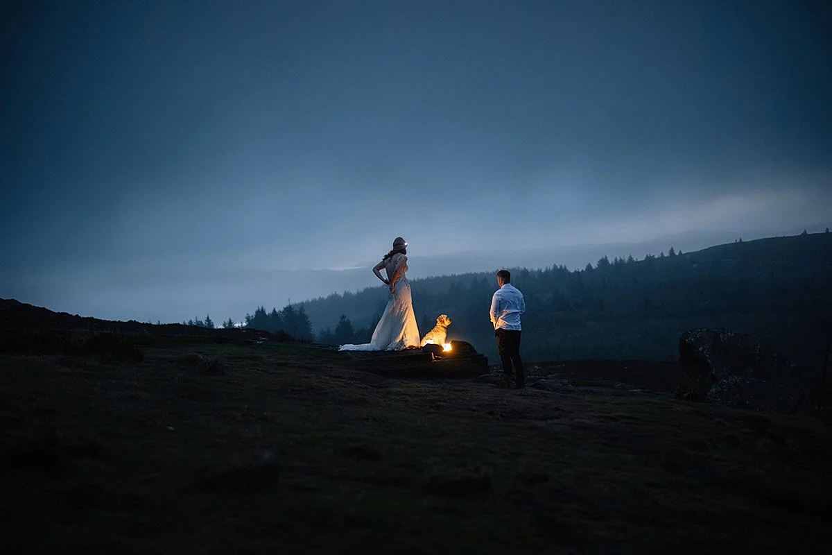 A couple with a dog standing on a hill under a dark sky, with distant mountains and a forest in the background.