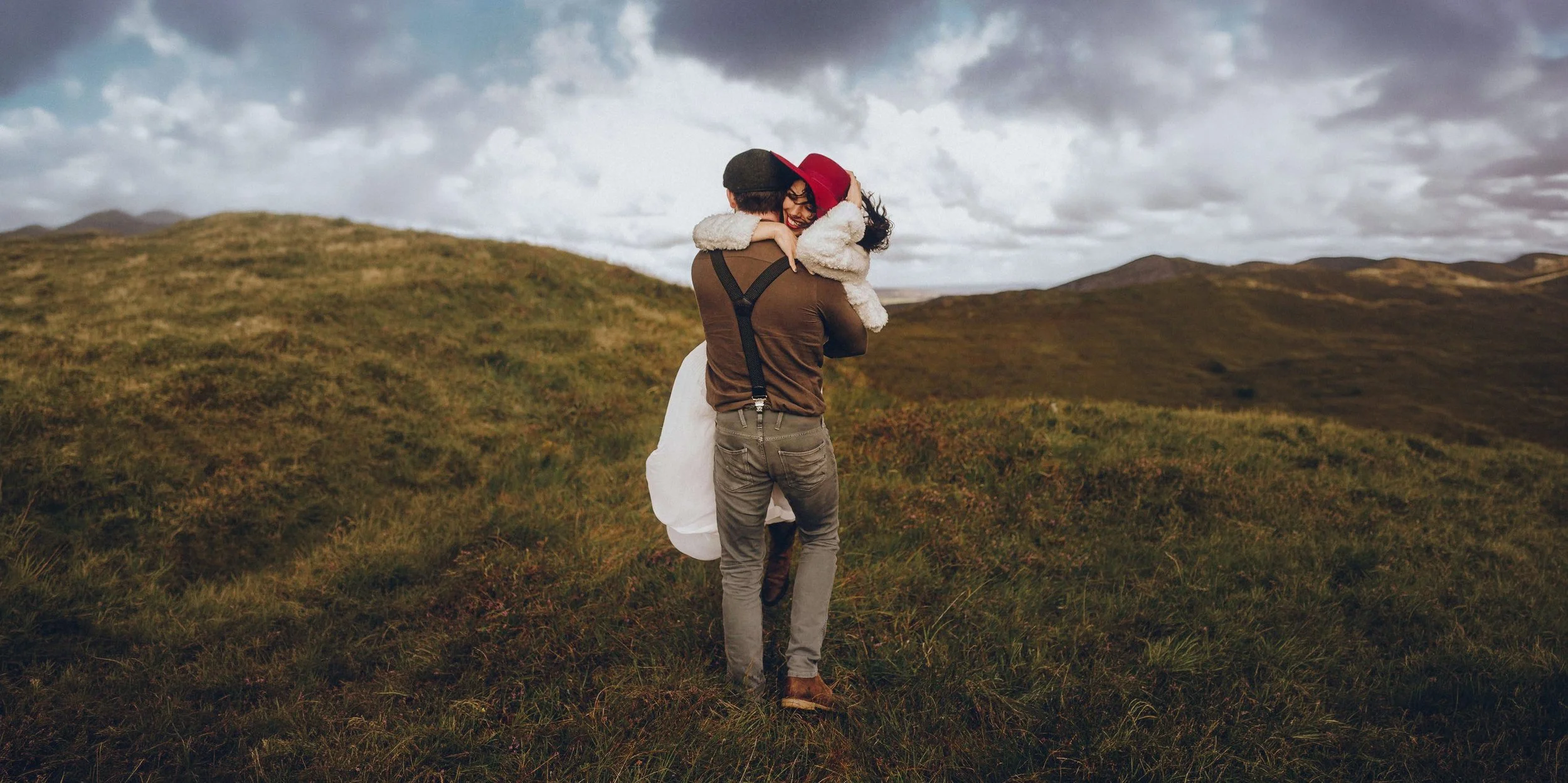 A man and woman embrace on a grassy hillside with cloudy skies in the background, with the man carrying the woman.