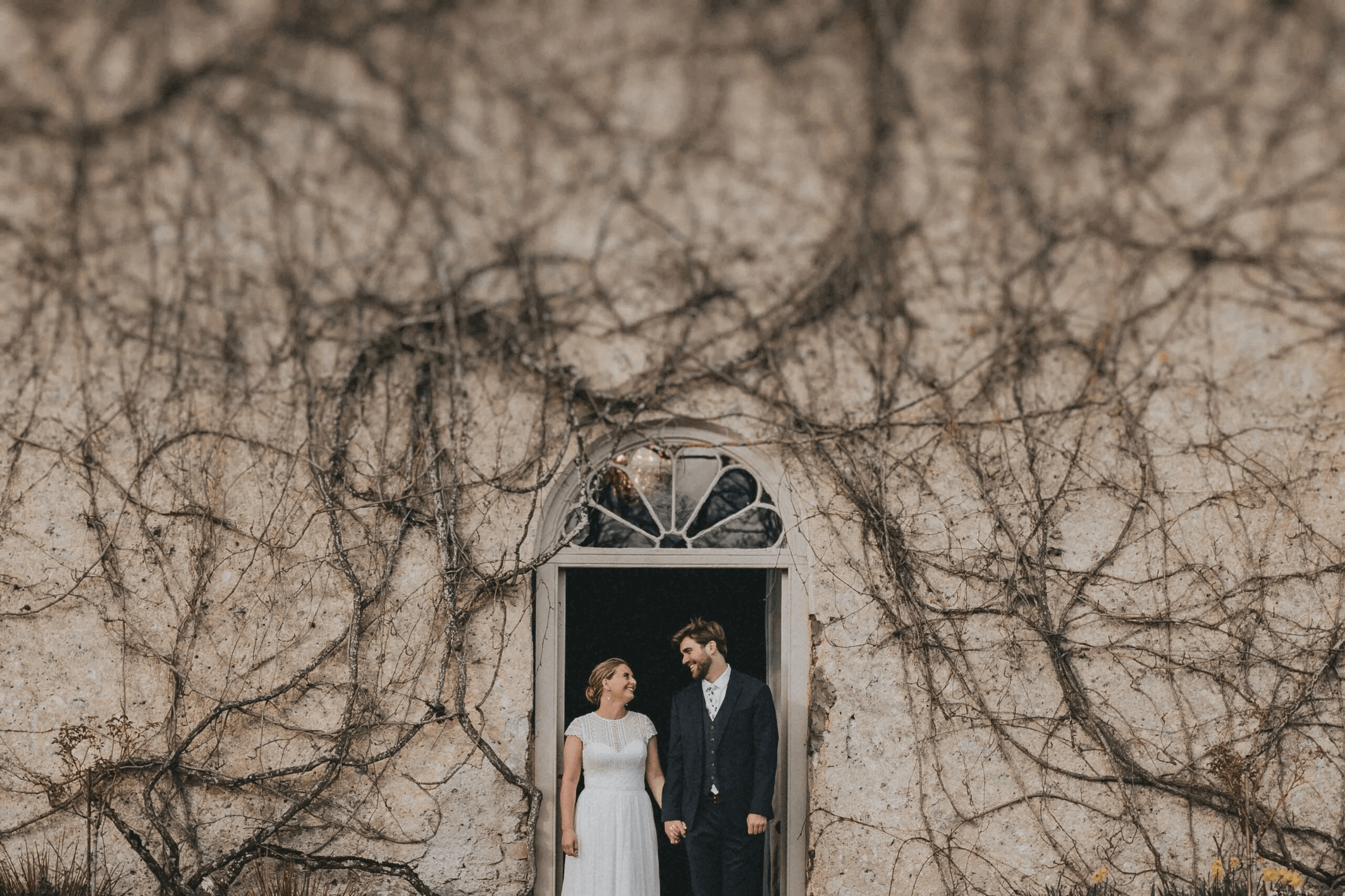 A couple in wedding attire standing in a doorway framed by a stone wall with climbing vines at Cloughjordan House during their wedding