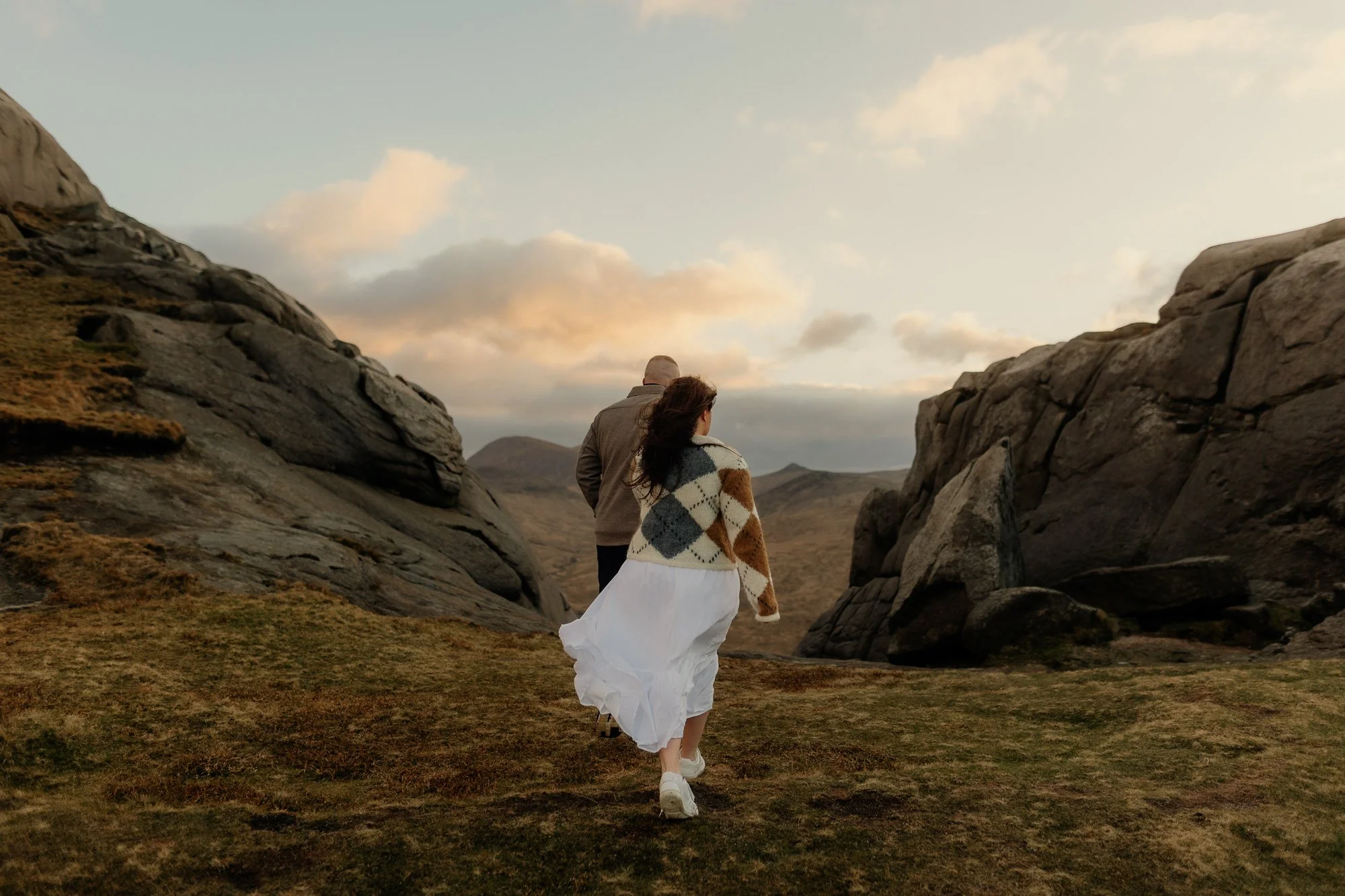 Couplewalk, a girl  in a white skirt and white sneakers walking through a rocky valley with large stone formations on each side, under a sky with scattered clouds.