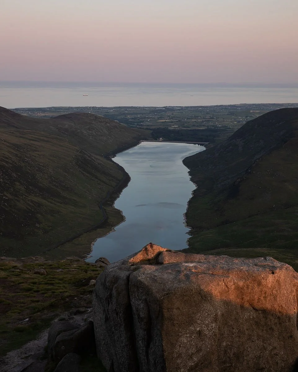 A scenic view of a narrow lake between two green hills, with a large rock in the foreground and a distant ocean in the background at sunset.