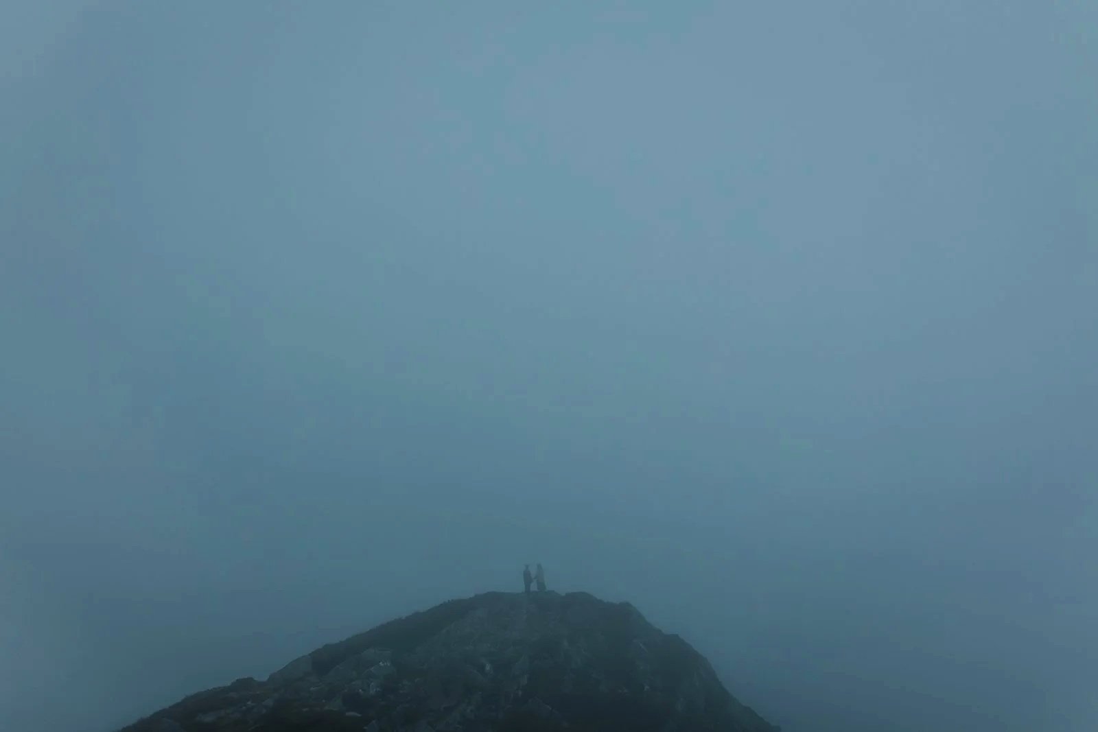 Two people standing on top of a mountain, silhouetted against a foggy, gray sky.