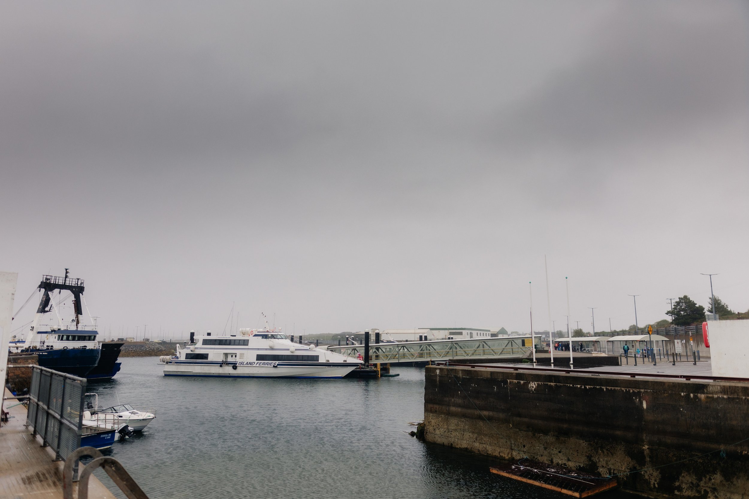 A marina with boats docked, including a boat labeled 'Island Ferries,' under an overcast sky with dark clouds, and a walkway along the water's edge with a few pedestrians.