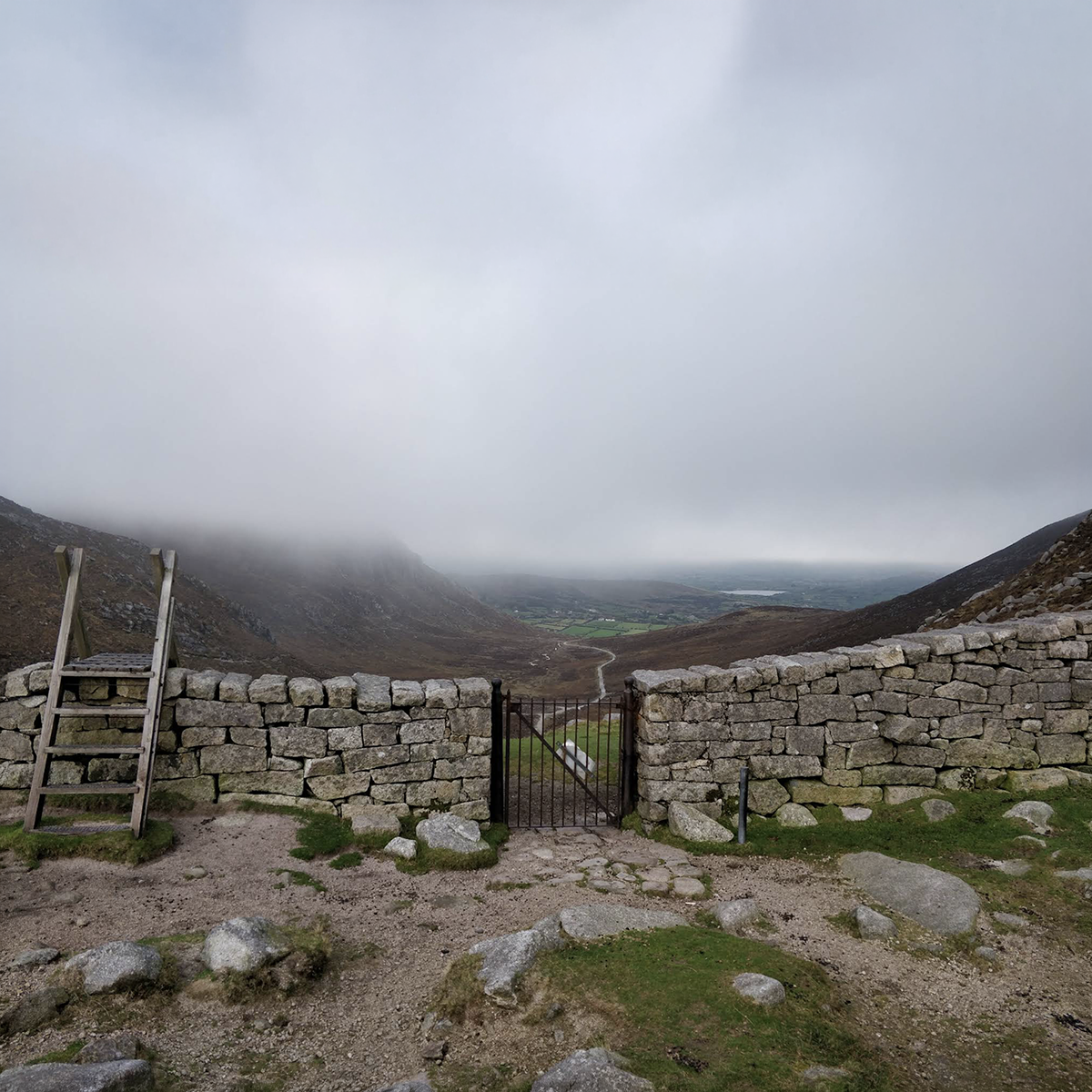 A stone wall with a metal gate in the center, leading to a hiking trail in a mountainous landscape under overcast sky, with low clouds covering the peaks and a view of distant fields and water bodies.