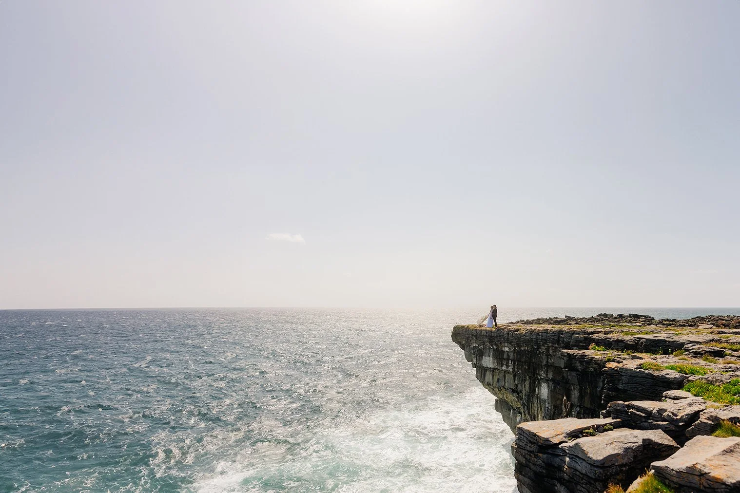 A couple standing on a rocky cliff over the ocean, with sunlight reflecting off the water, celebrating their wedding.