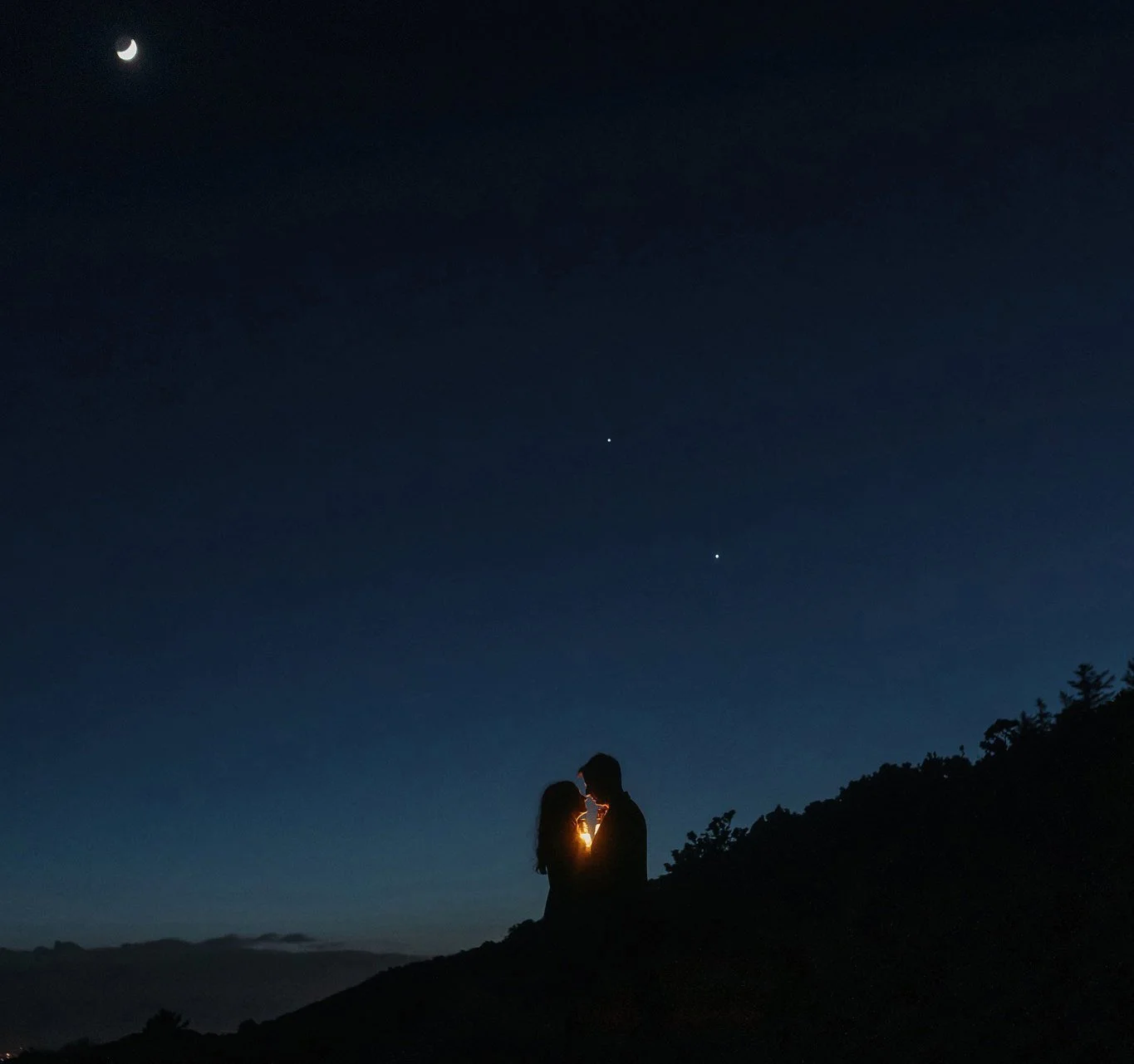 Silhouette of a couple embracing on a hillside at night under a starry sky and crescent moon during their adventure couple shoot  in Ireland.