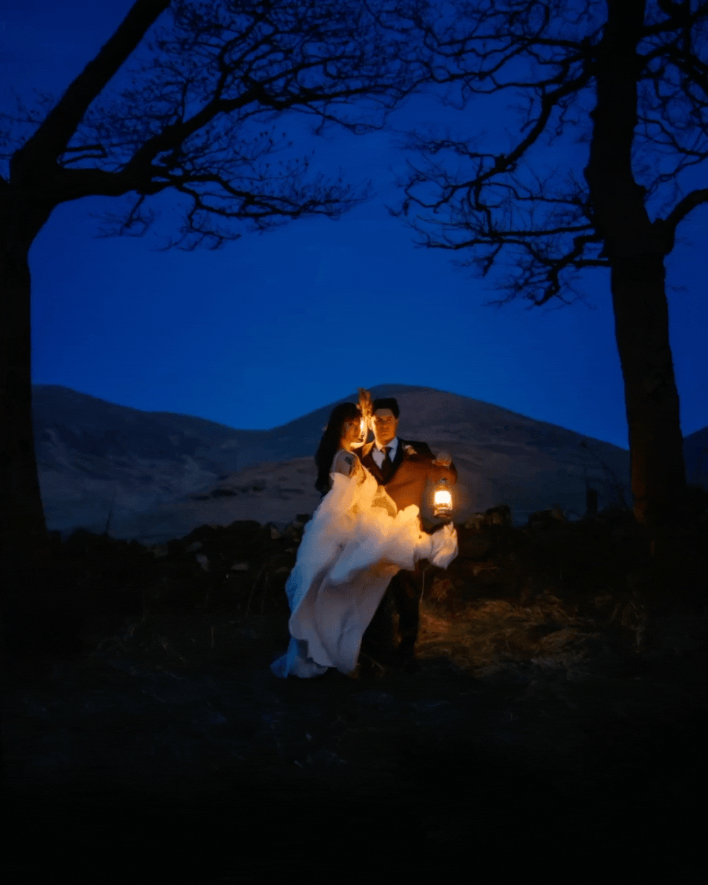 A couple dressed in wedding attire standing outdoors at night, illuminated by a lantern they are holding, with a dark blue sky, barren trees, and mountains in the background.