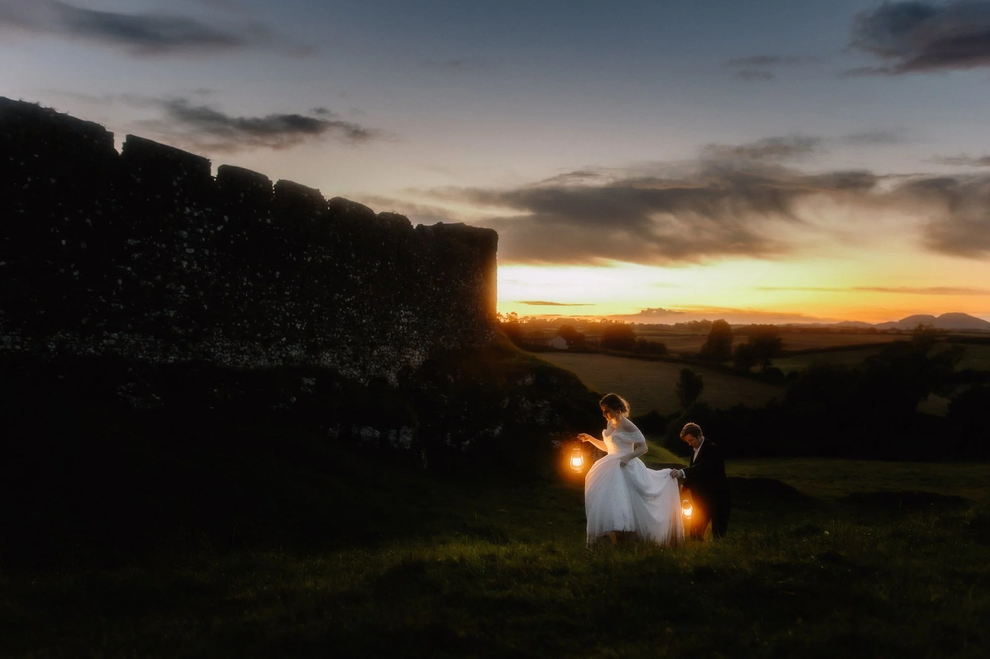 A bride and groom holding lanterns at sunset in a rural landscape with old stone structures and a cloudy sky.