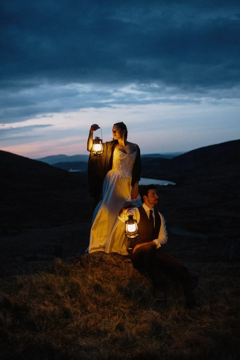 A man and a woman in period costumes holding lanterns outdoors during dusk, with hills and a river in the background.
