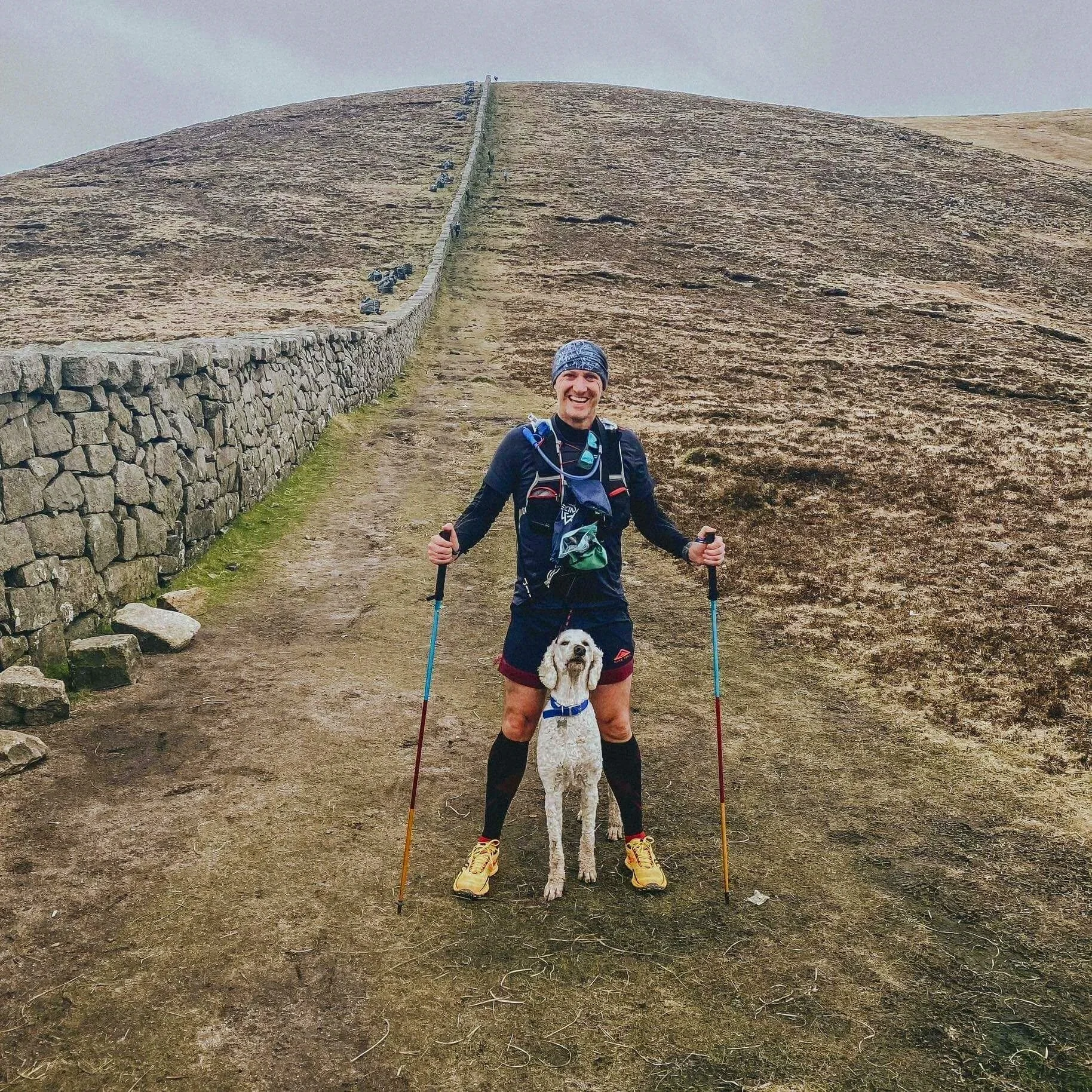 A man smiling while standing on a dirt trail, holding hiking poles, with a golden retriever dog. They are on a hill with a stone wall on the side, and a steep incline of grass or moss behind them.